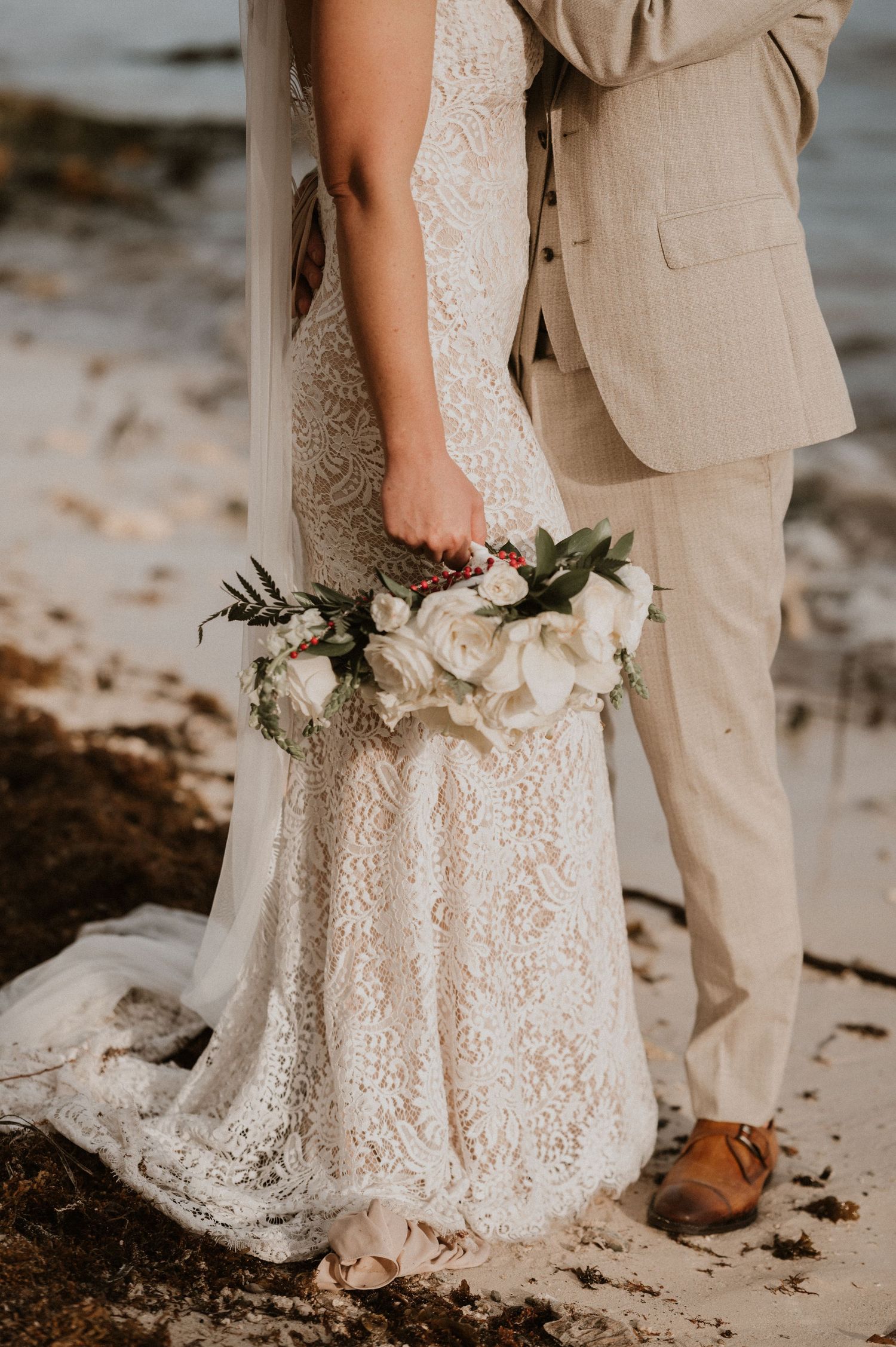Close-up of white lace wedding dress and beige suit on beach.