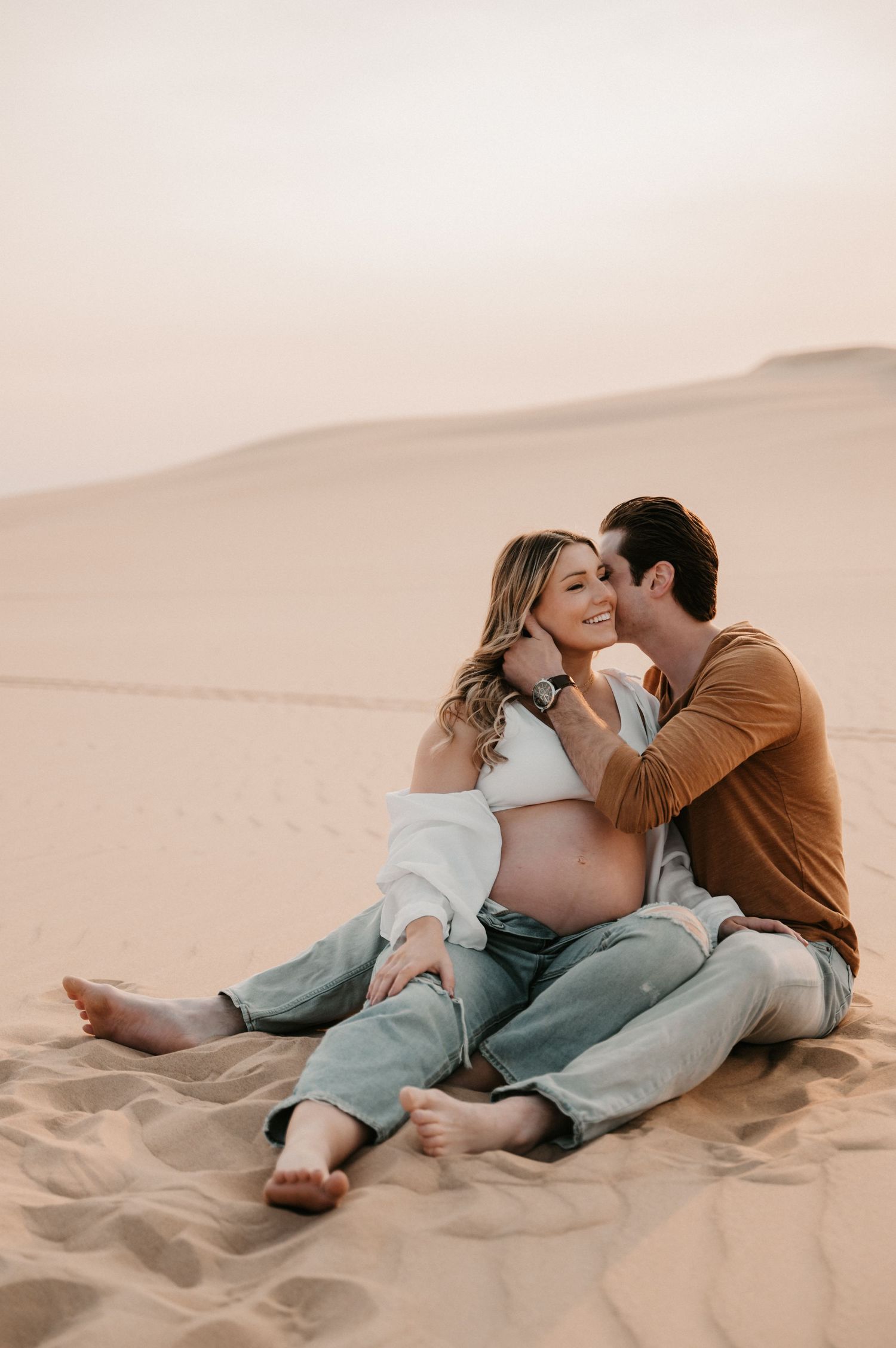 Maternity photo session captures intimate moment between couple on desert dunes at sunset.