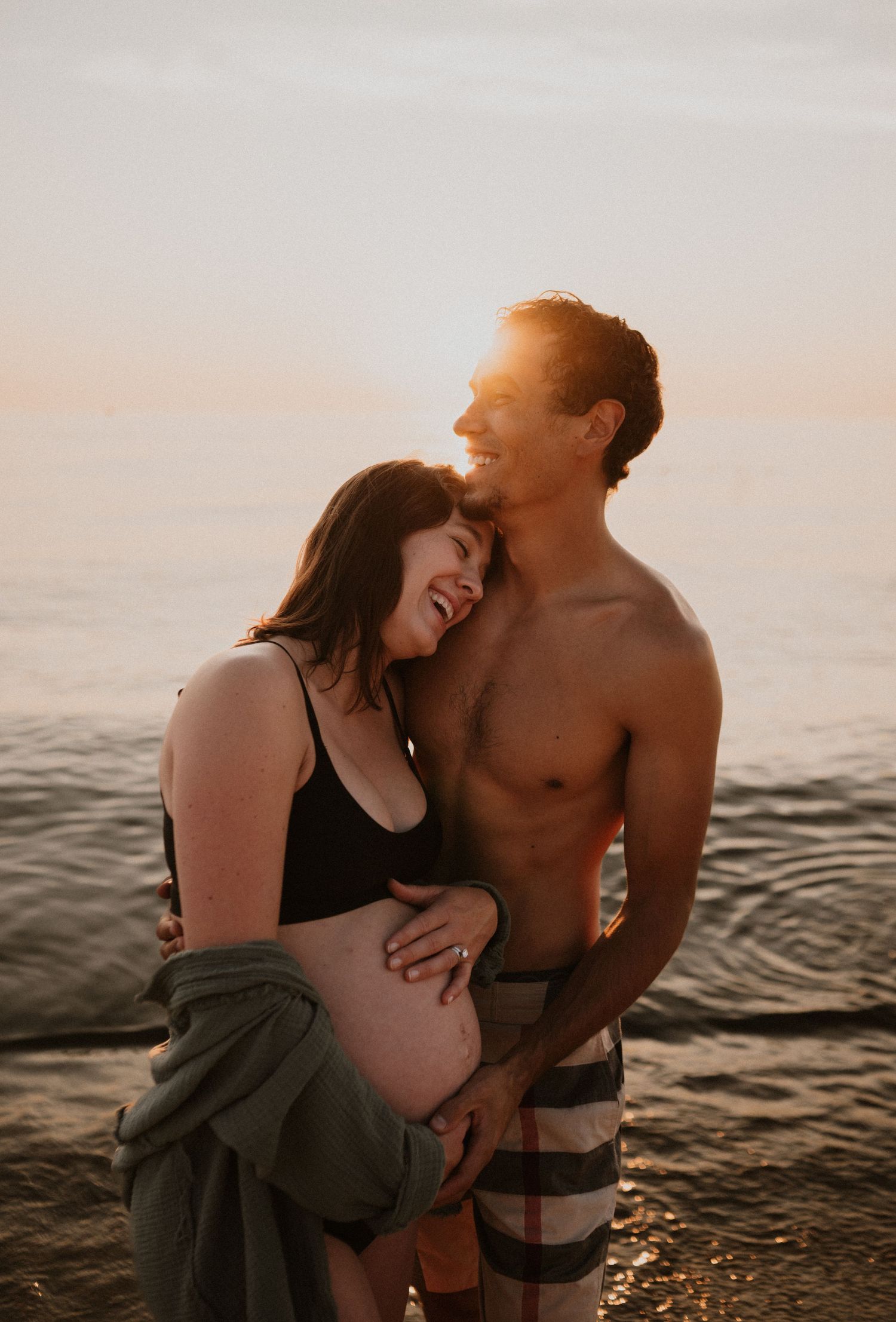 Loving maternity moment captured on beach with sunset glow and ocean waves.