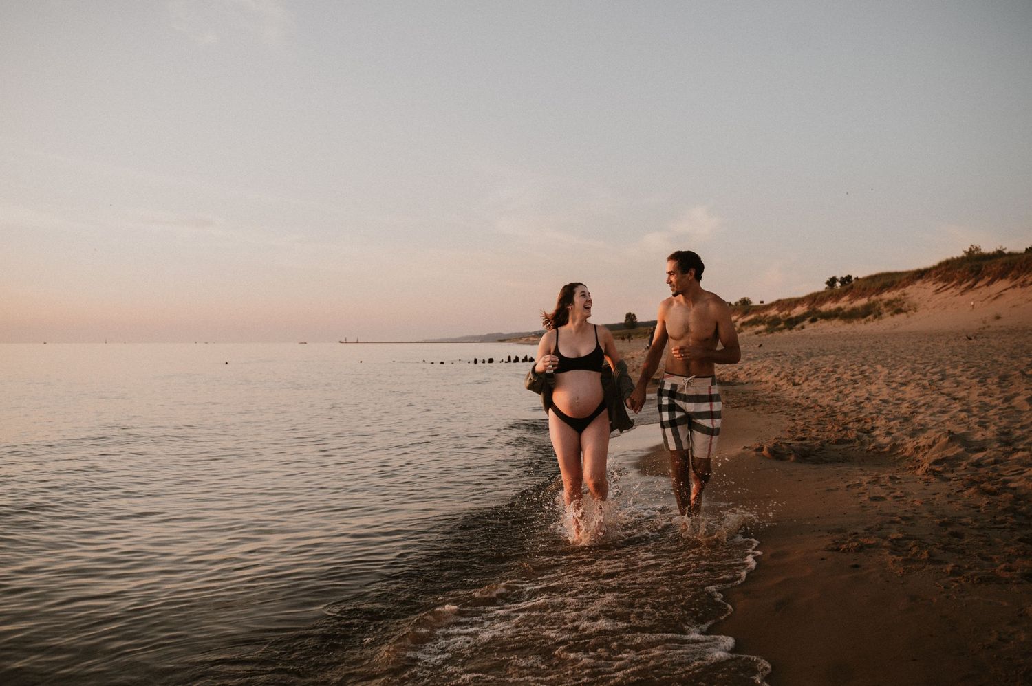 Expectant couple walking together along the shoreline during golden hour sunset.