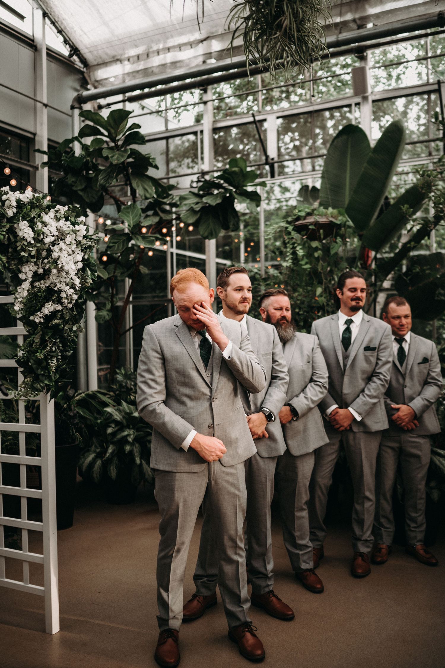 Wedding party in gray suits standing together in a modern greenhouse with lush plants.