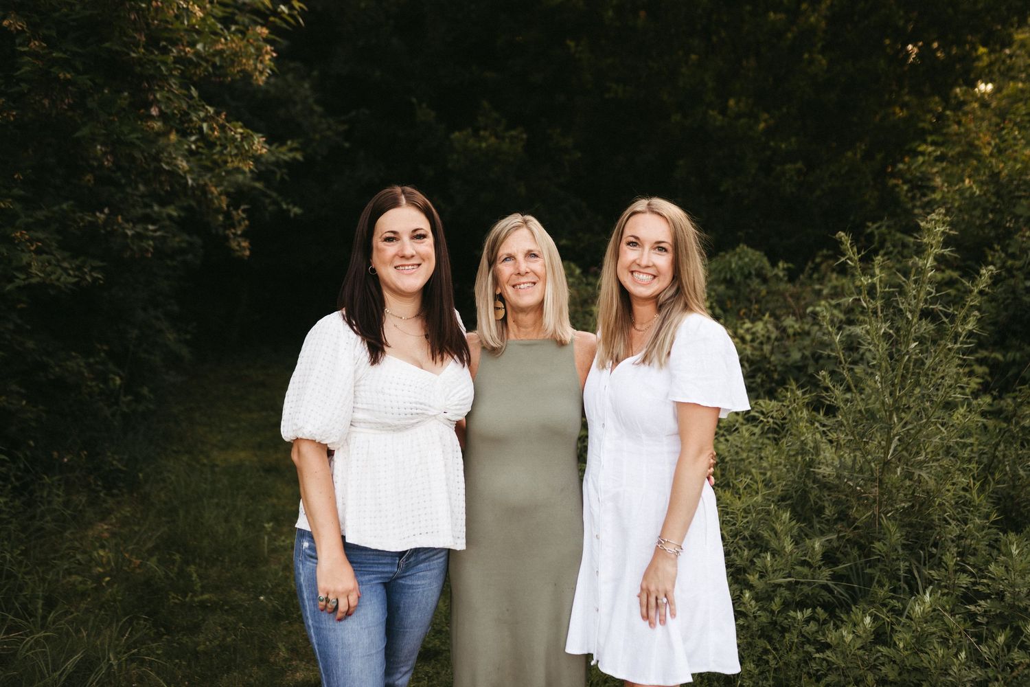 Three women pose together in neutral colored outfits against green foliage.