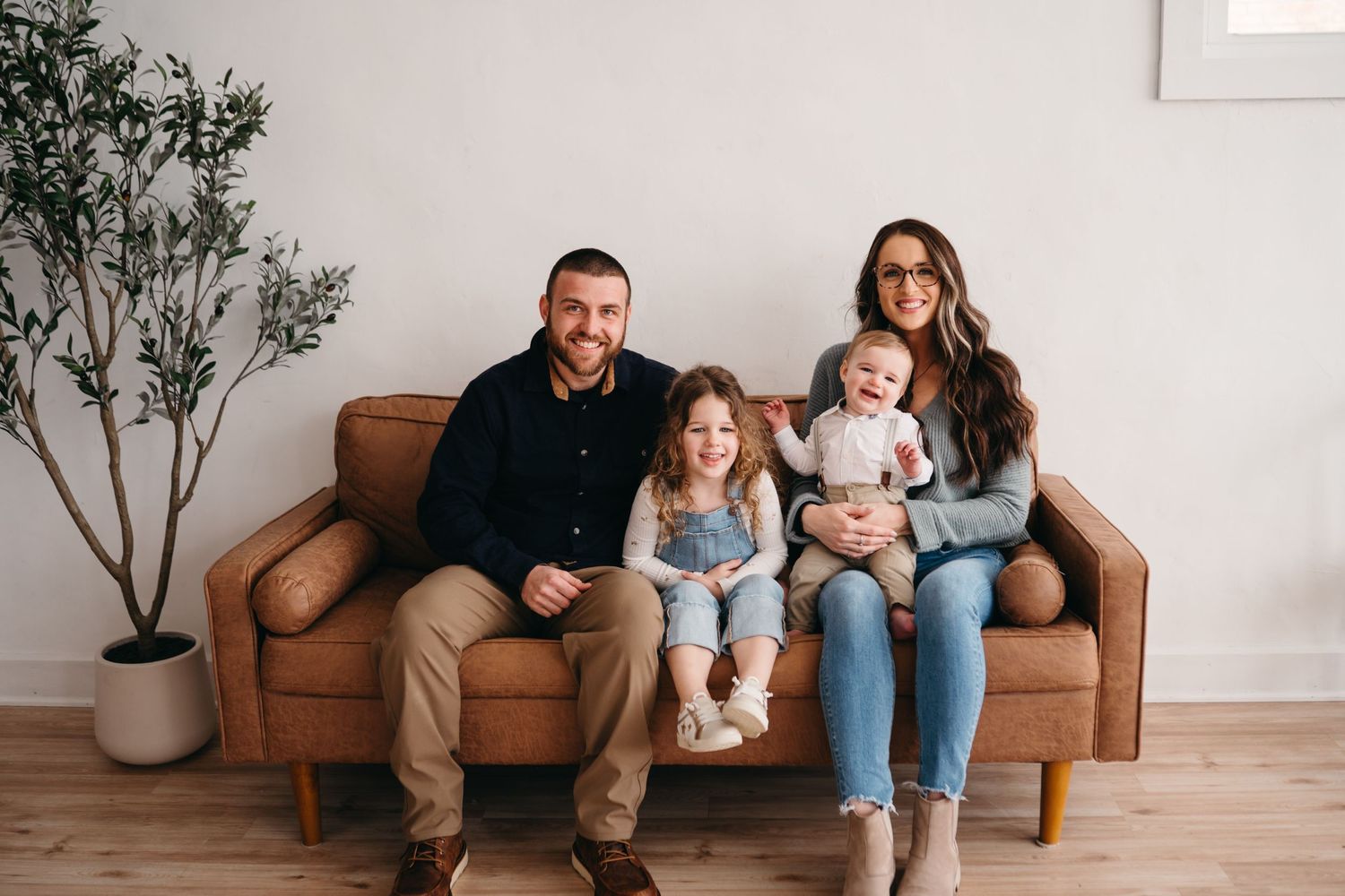 A family of four sits together on a brown leather couch in a minimalist living room with a potted plant.