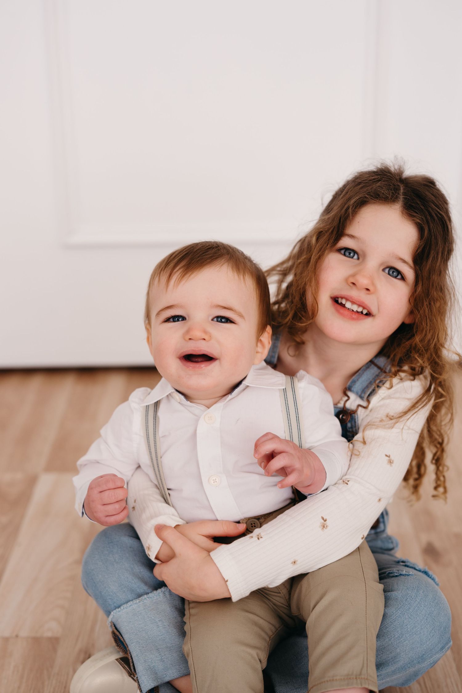 Two children pose together on hardwood flooring while sharing laughs and hugs.