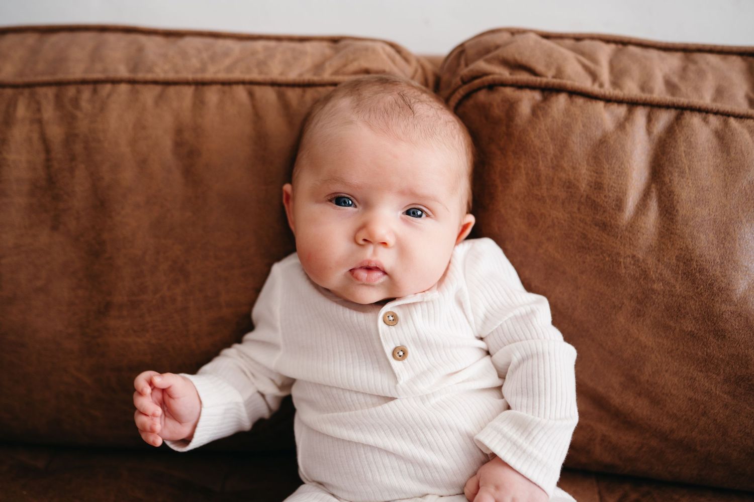 Infant in cream colored knit outfit rests against brown leather sofa cushions.