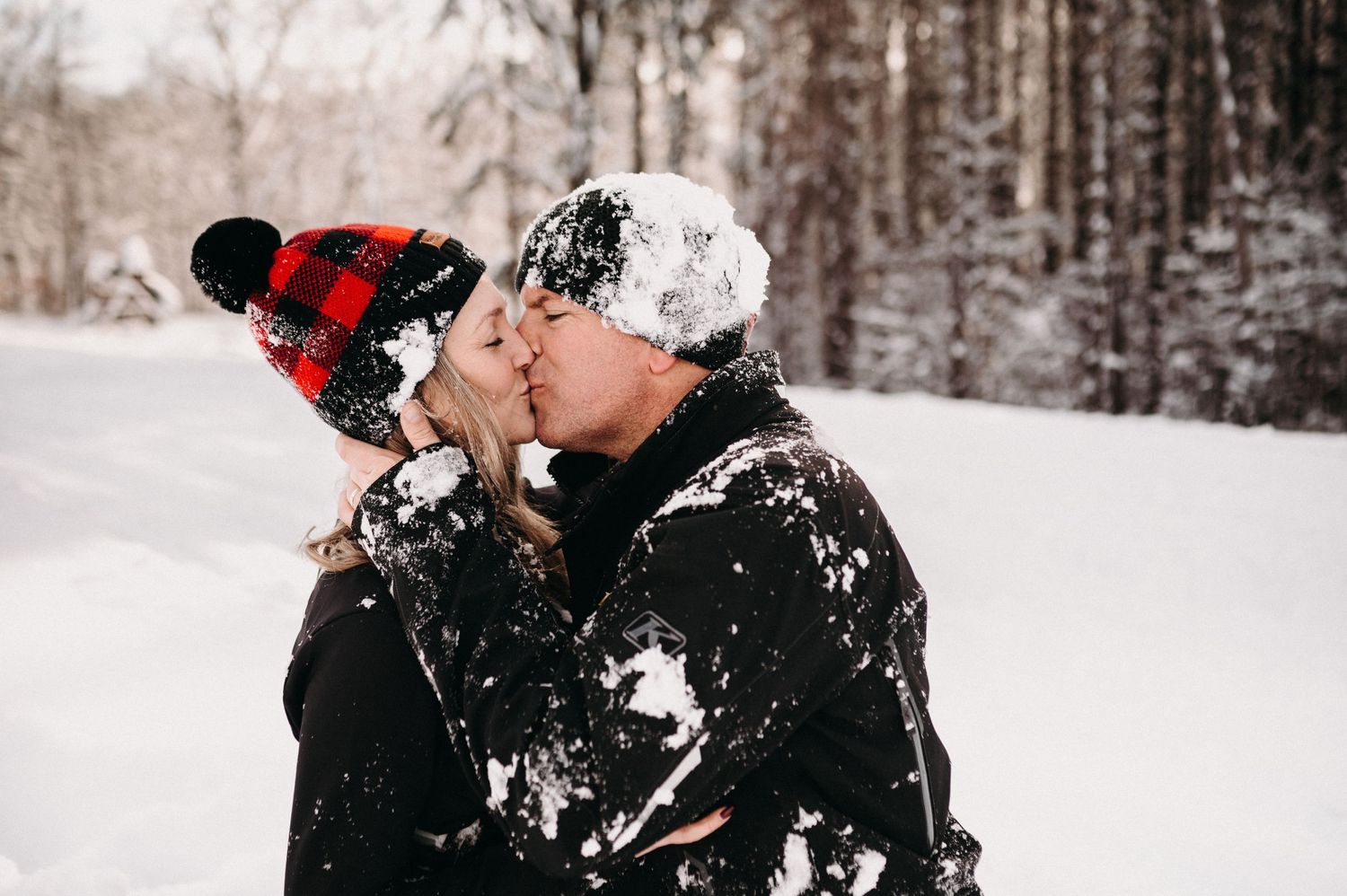 A romantic winter scene with two people in black winter gear and red plaid beanies sharing a moment.