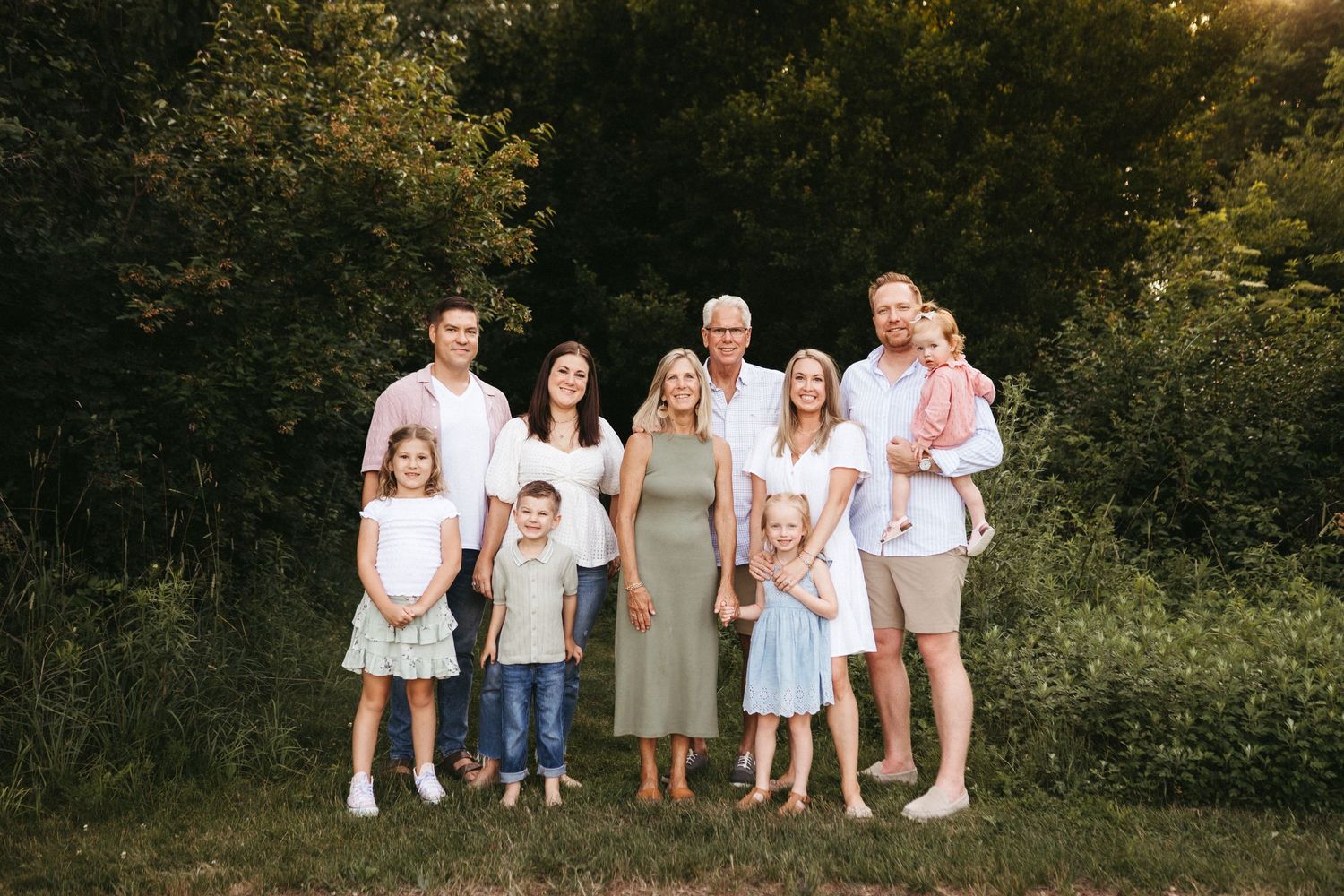Extended family gathering for outdoor photos against a green leafy backdrop at sunset.