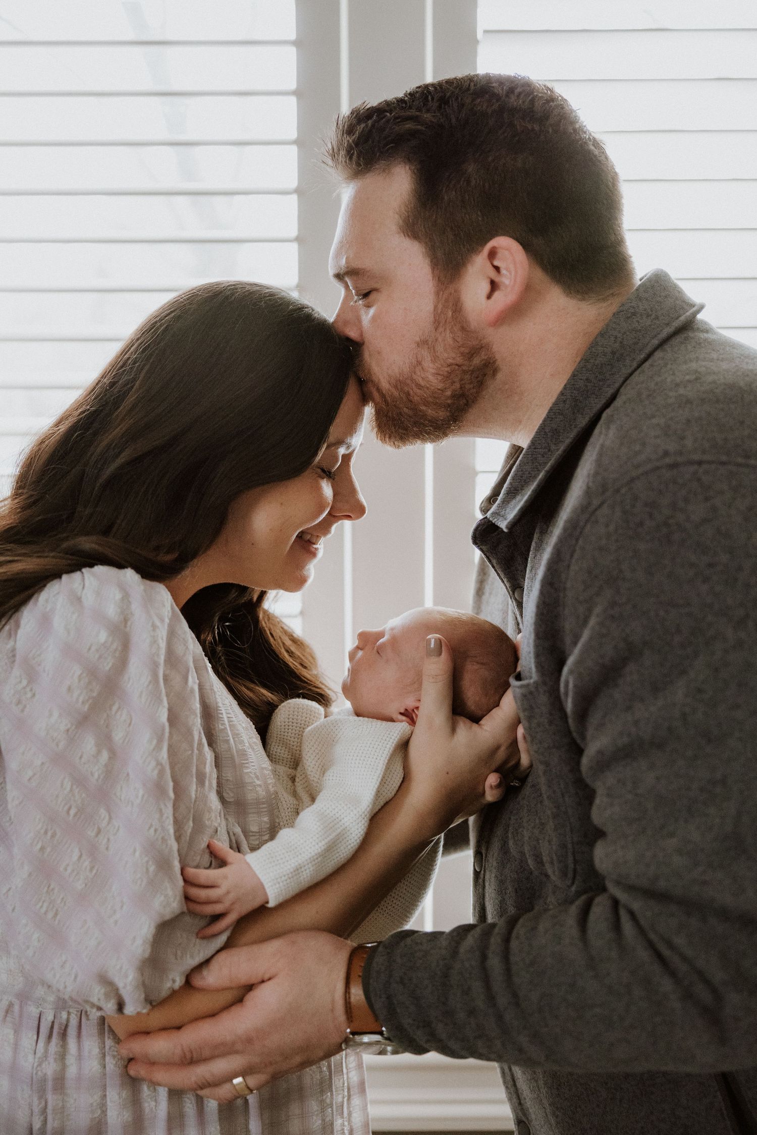 Sweet family moment sharing kisses with baby near a bright window.