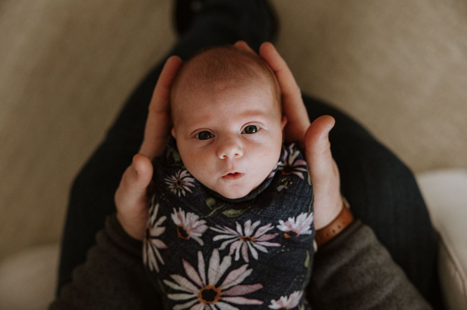Newborn baby wearing floral print outfit being gently held in natural light setting.