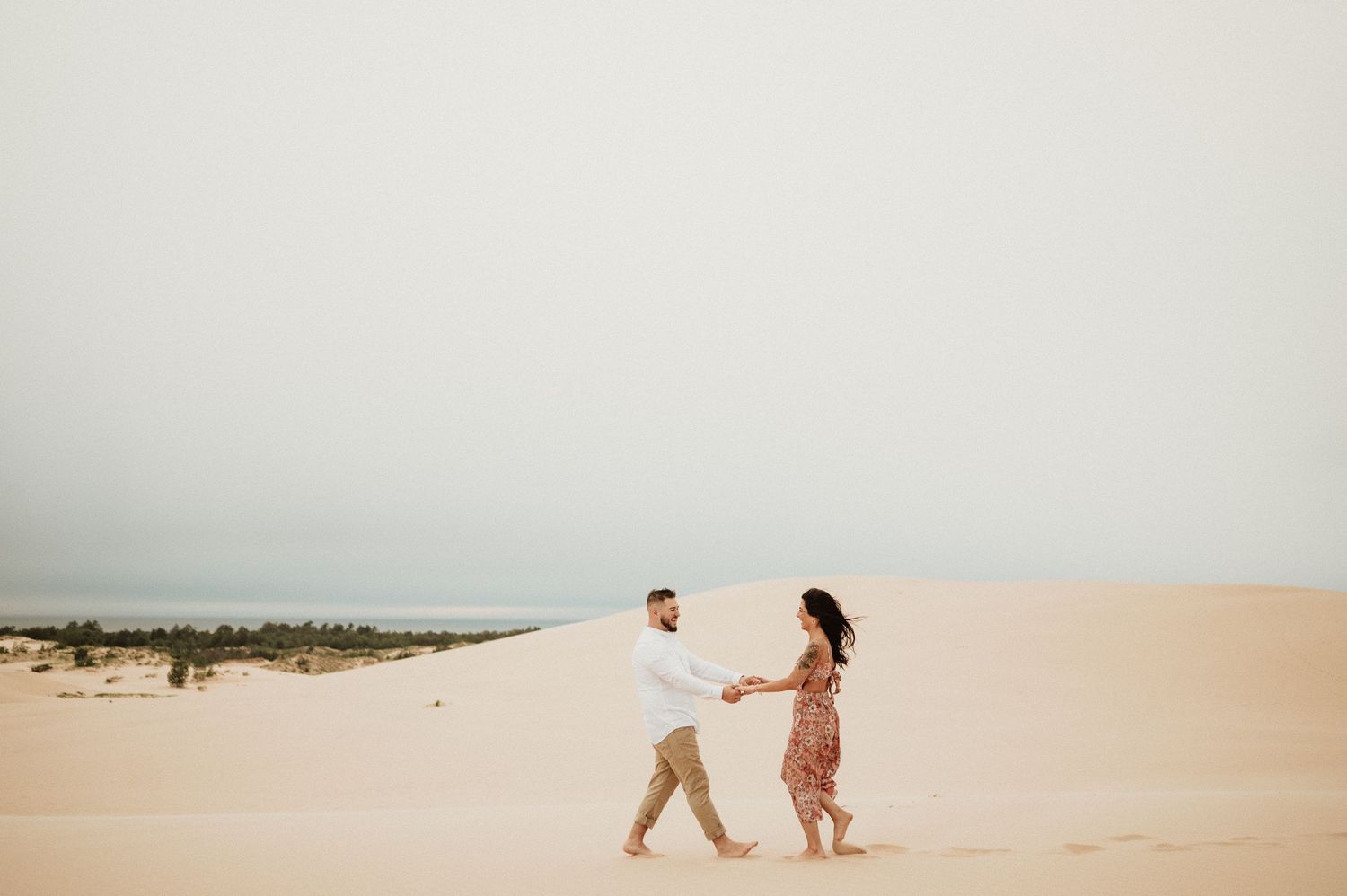 A couple holding hands and walking together across sweeping sand dunes in matching light-colored outfits.
