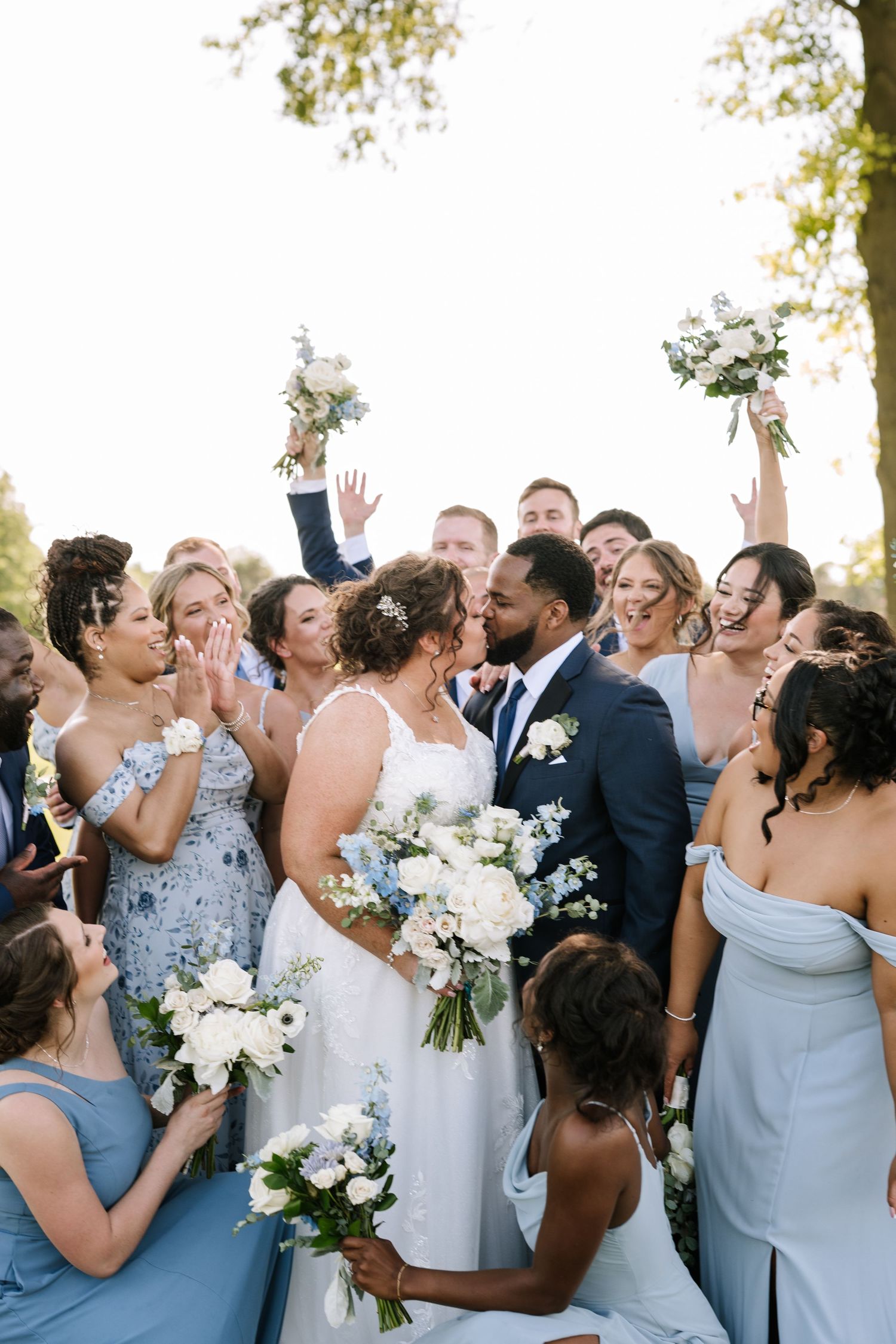 A wedding party in light blue and navy attire celebrates joyfully together outdoors during a summer ceremony.