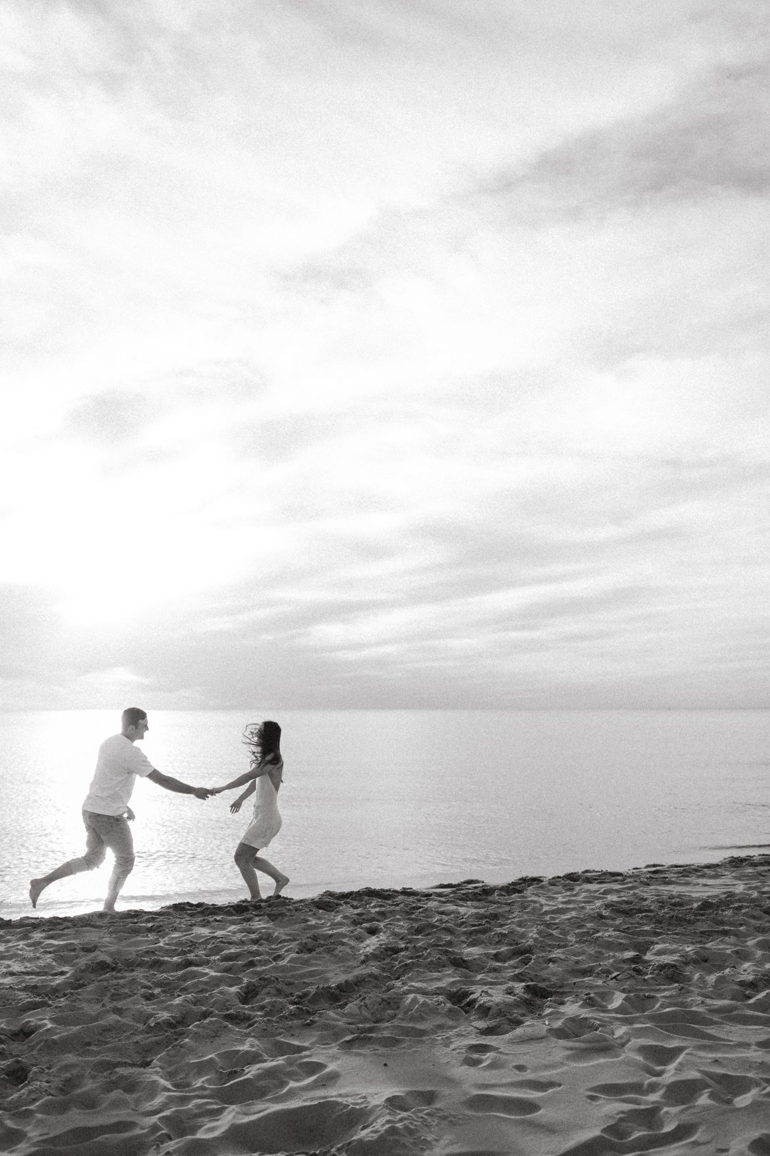 Black and white photo of a couple running hand in hand along a sandy beach at sunset.