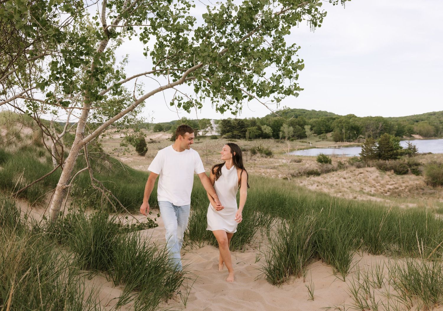 Happy couple walking barefoot on sandy dunes surrounded by lush greenery and a serene lake.