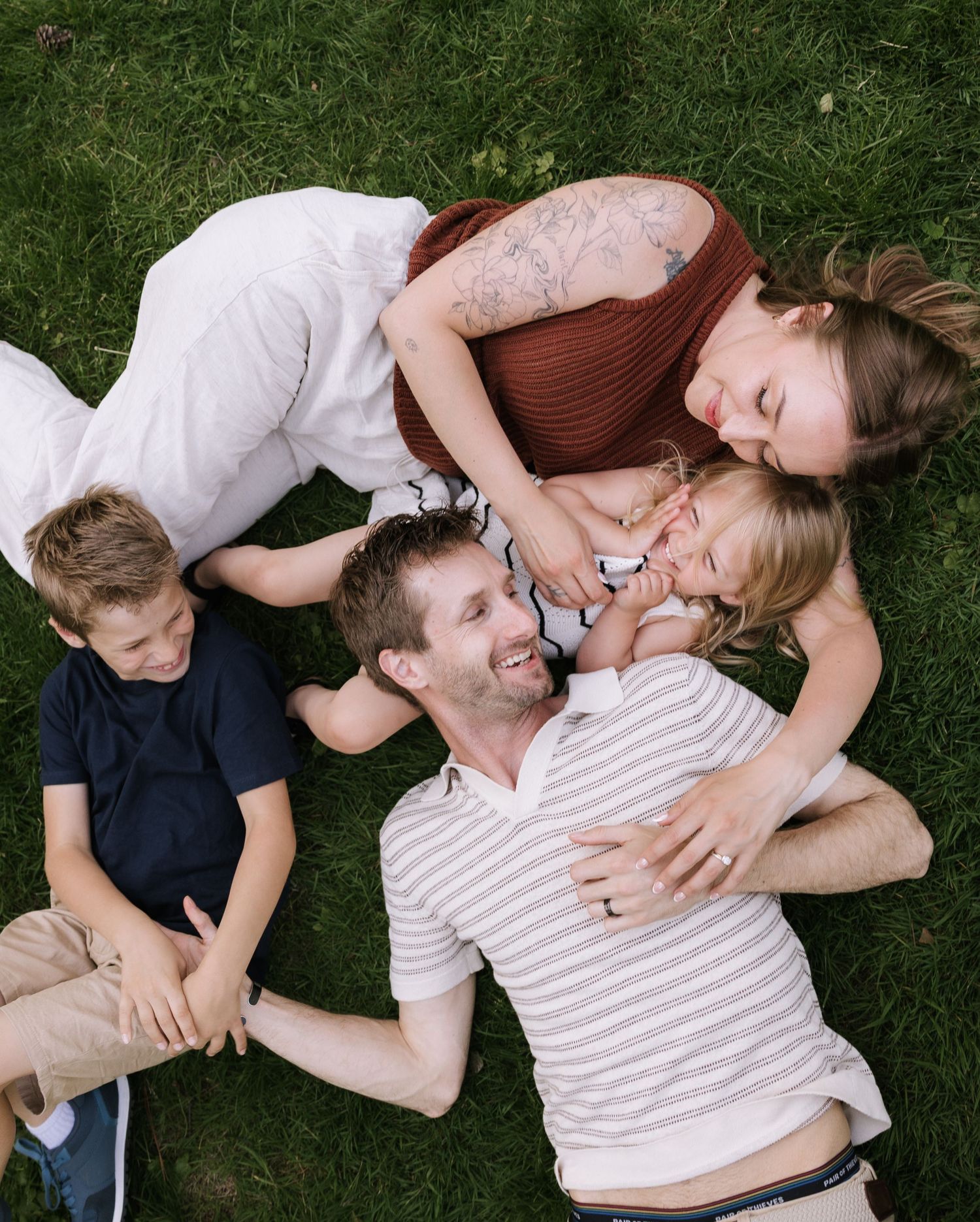 A happy family of four laughs together while lying on green grass in a park.