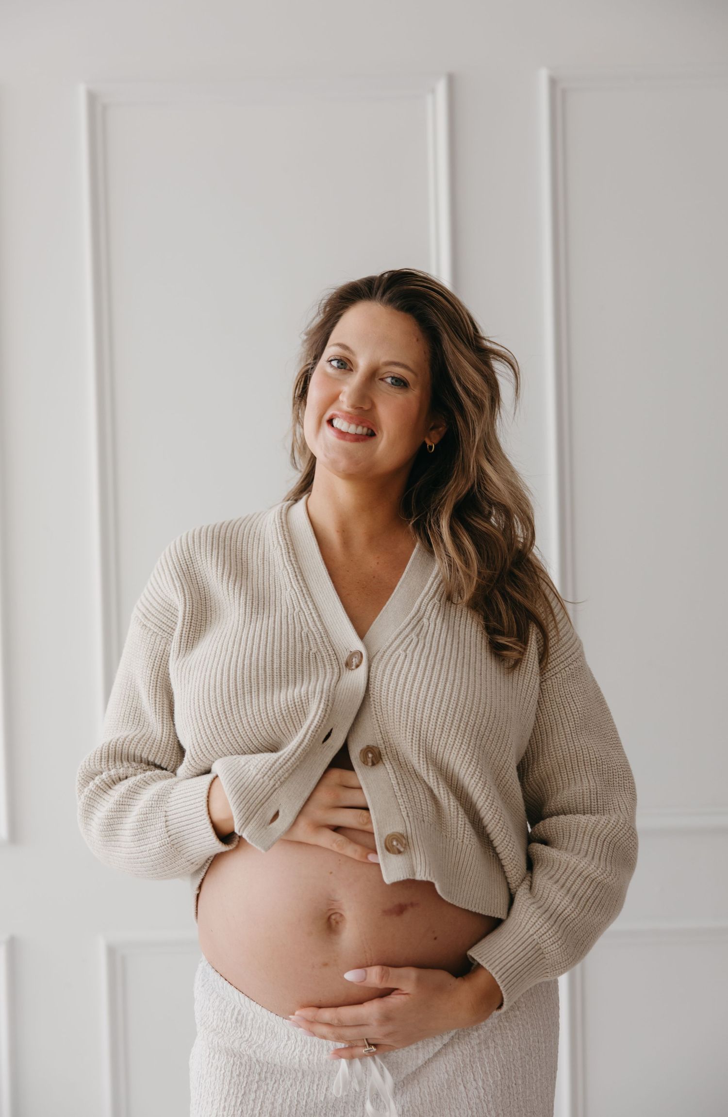 Smiling pregnant woman in beige ribbed cardigan cradling baby bump against white wall.
