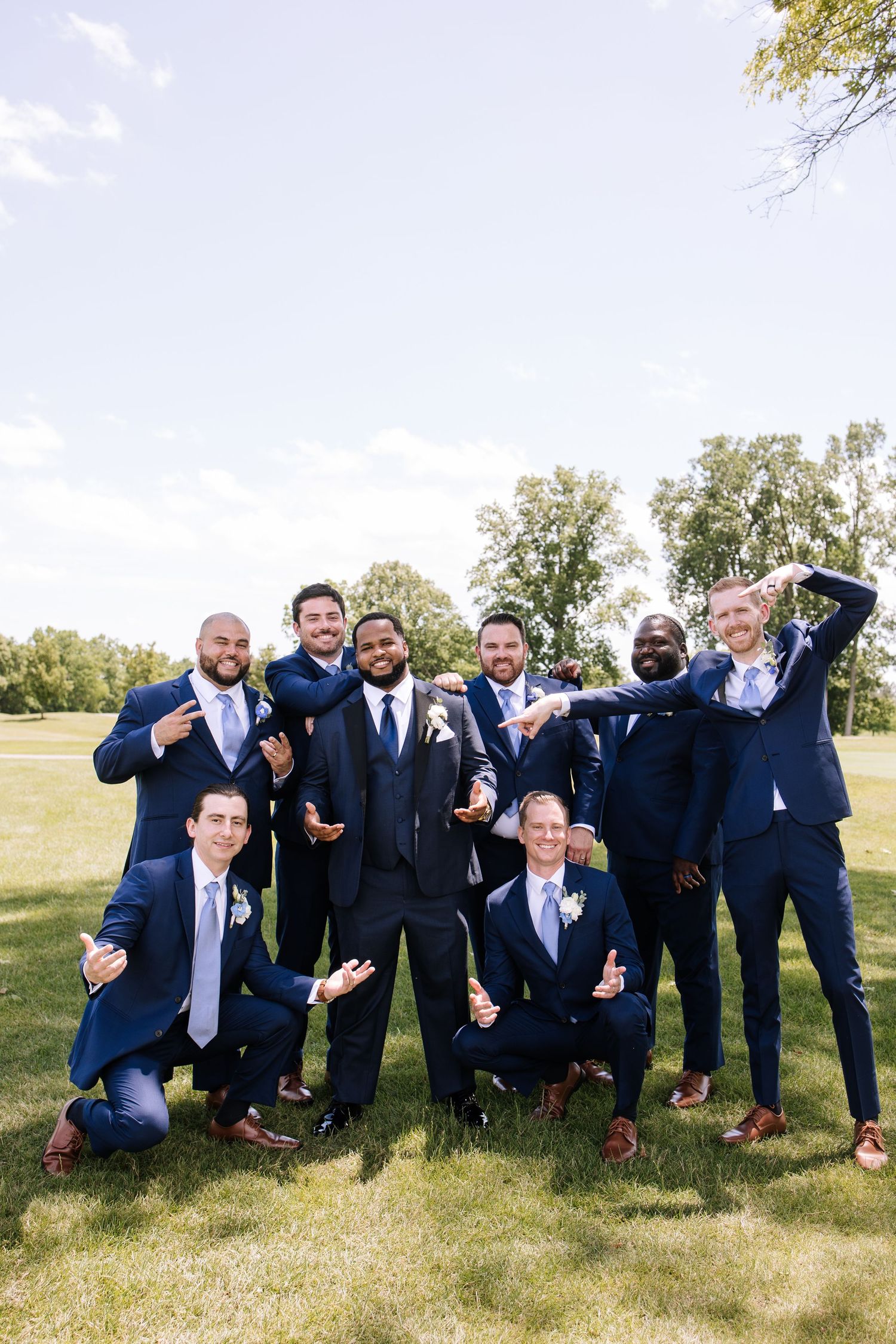 Groomsmen in navy suits posing playfully on grass for wedding photos.
