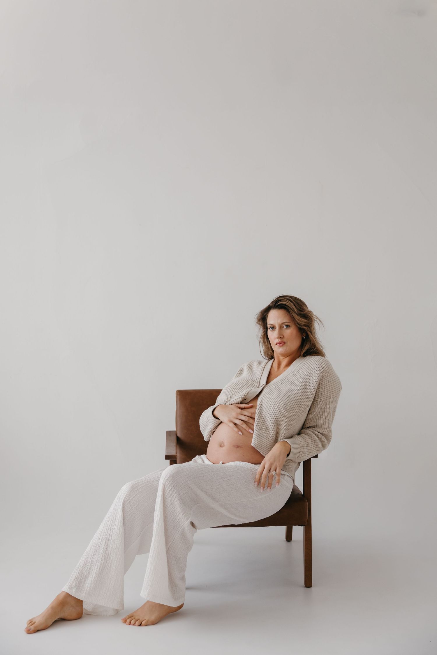 Pregnant woman in beige cardigan and white pants sits in wooden chair, cradling bare baby bump in minimalist studio.