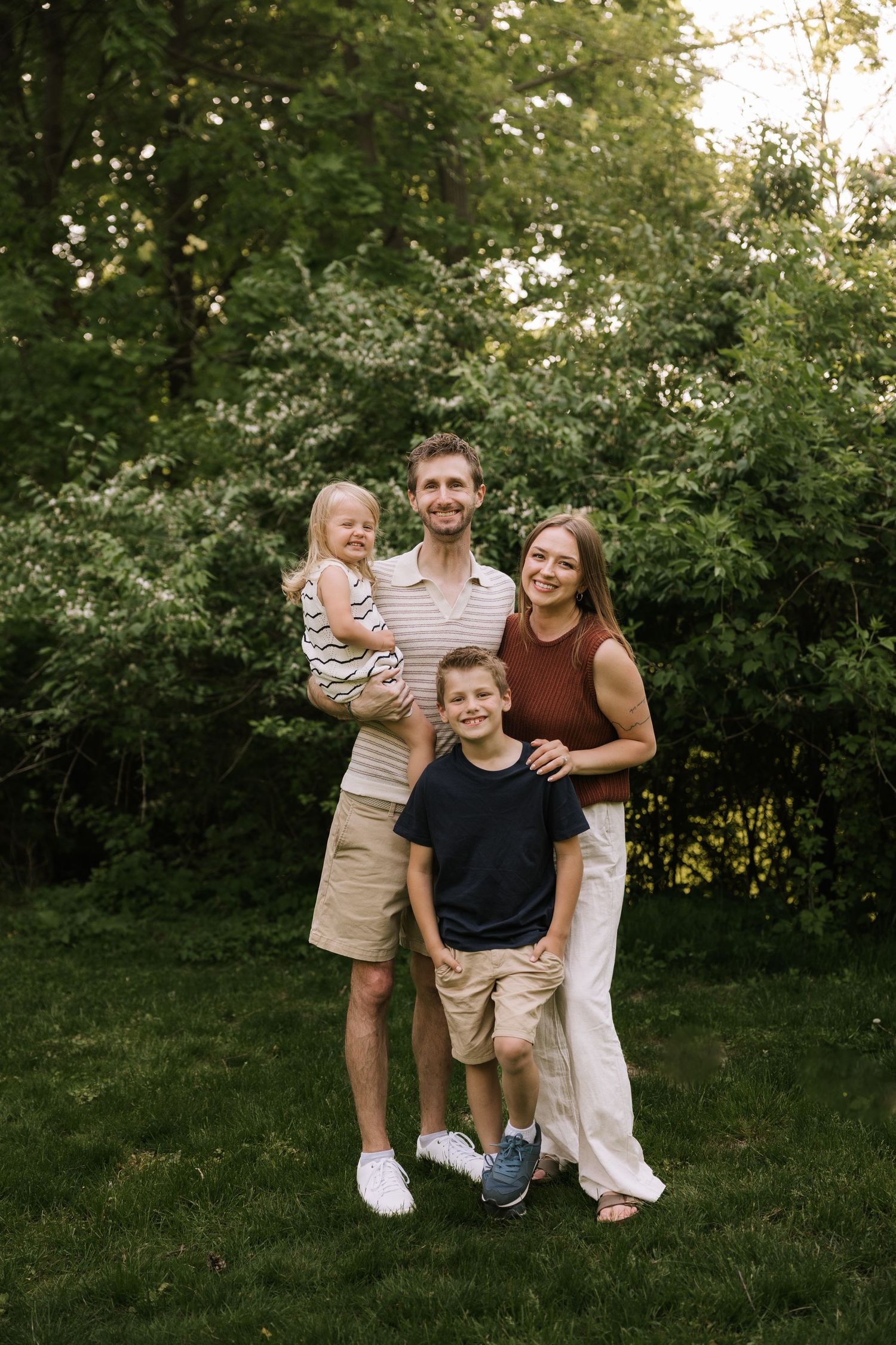 A happy family of four poses outdoors on green grass surrounded by lush trees during golden hour.