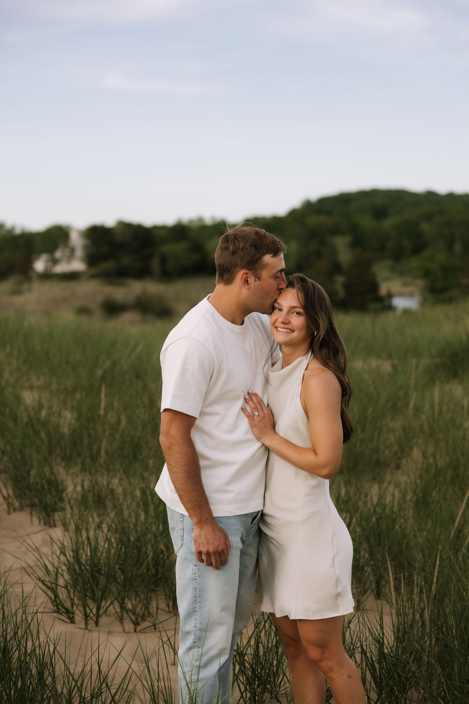 Couple embracing in sandy dunes, man kissing woman's temple, both wearing white, green grass background.