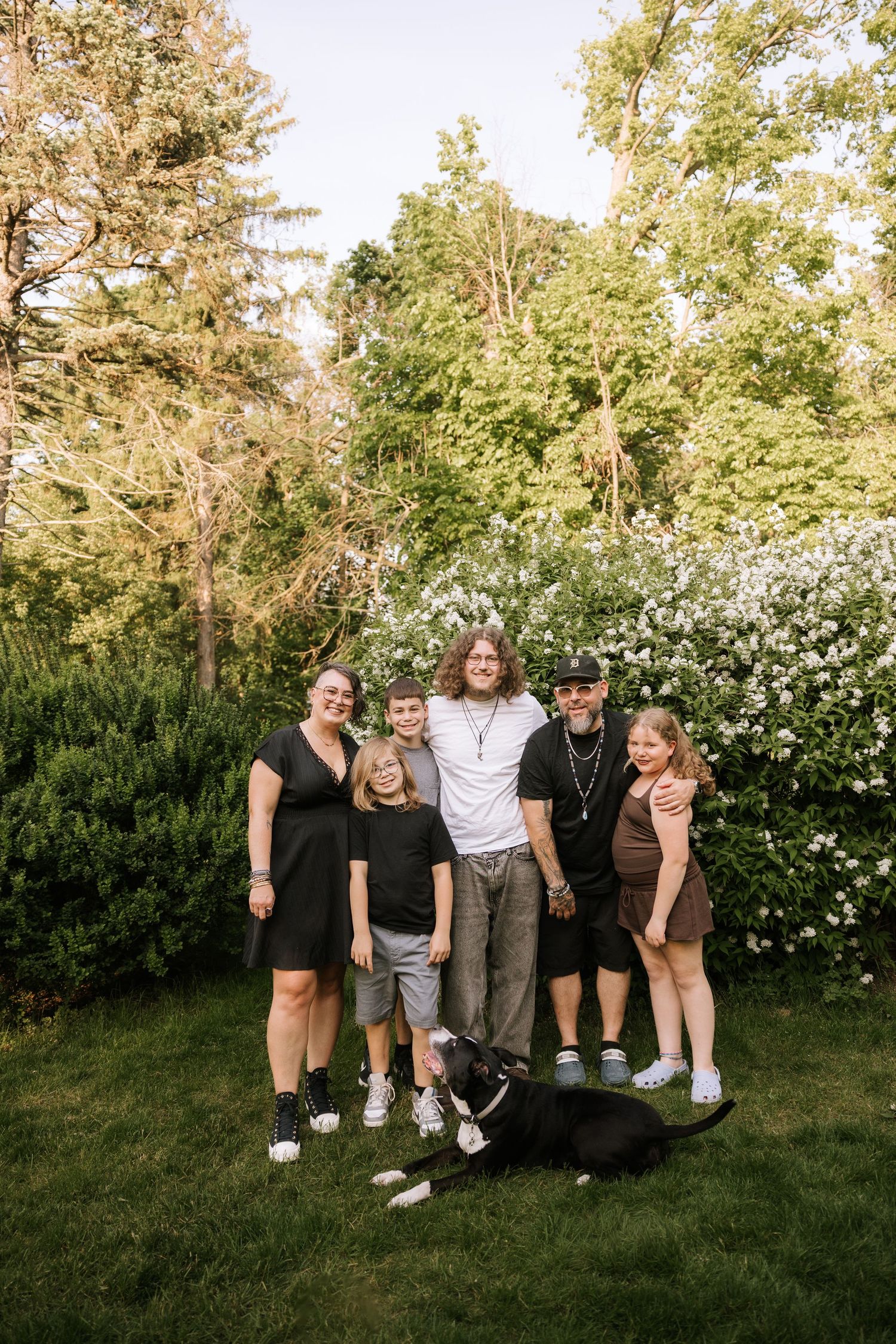 Family of five posing with their black Boxer dog in a garden with white flowering bushes.
