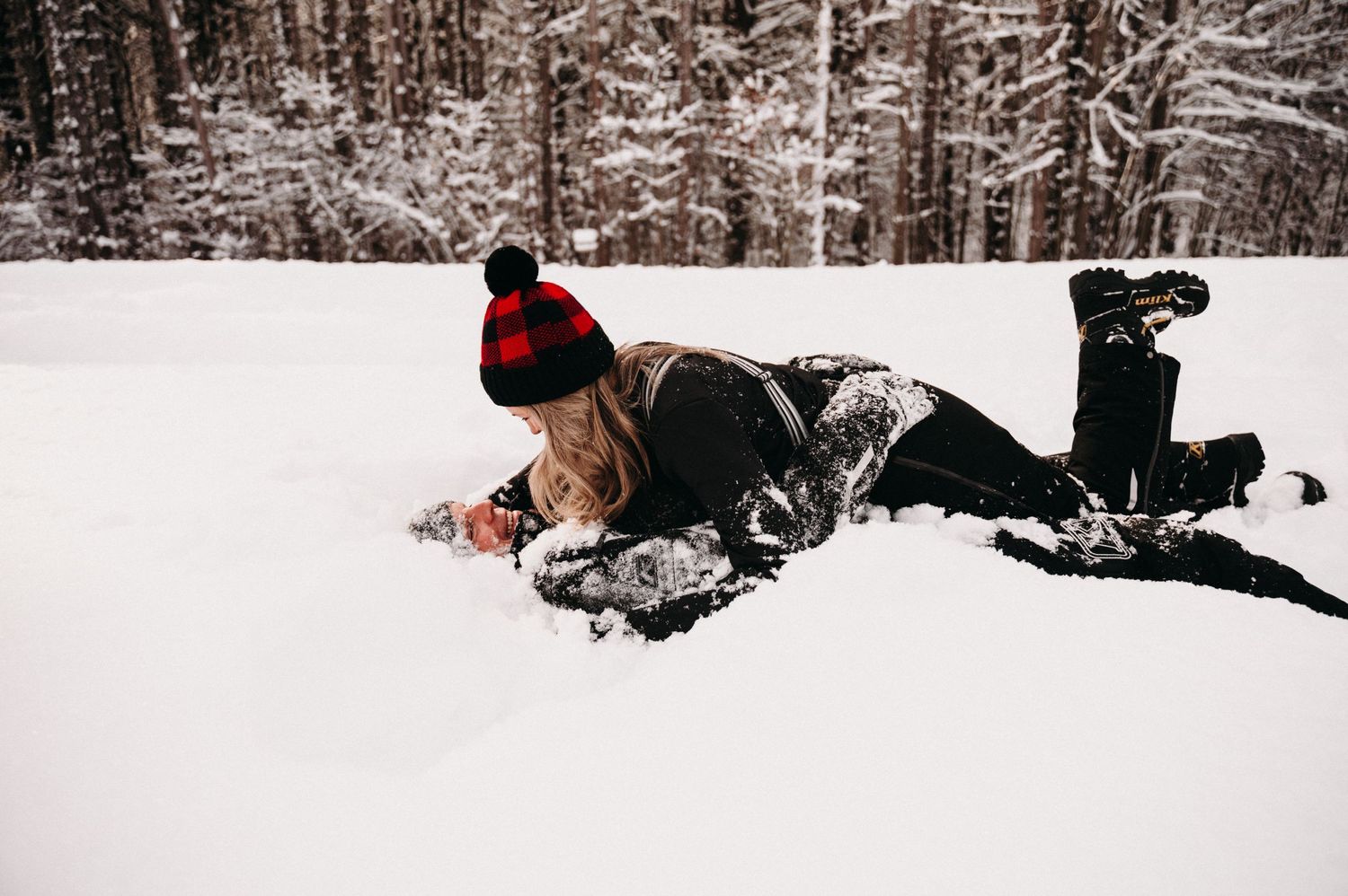Person in winter clothing lying in deep snow with snowy forest backdrop.