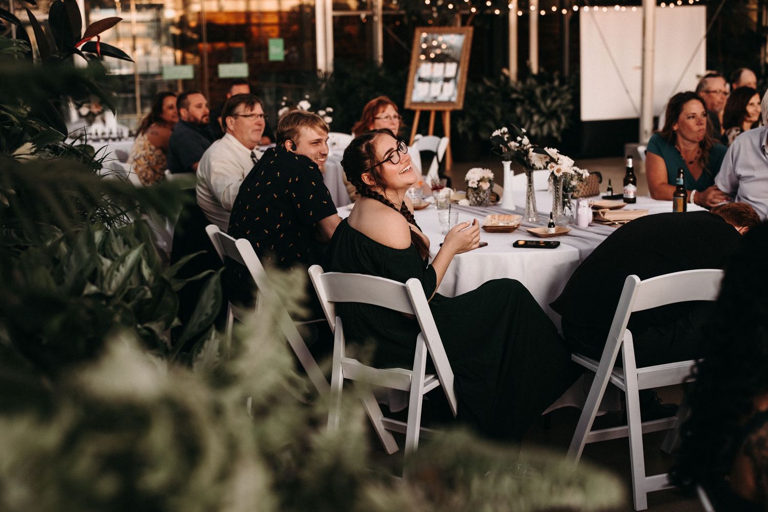 Wedding reception guests seated at white tables in a greenhouse with lush greenery.