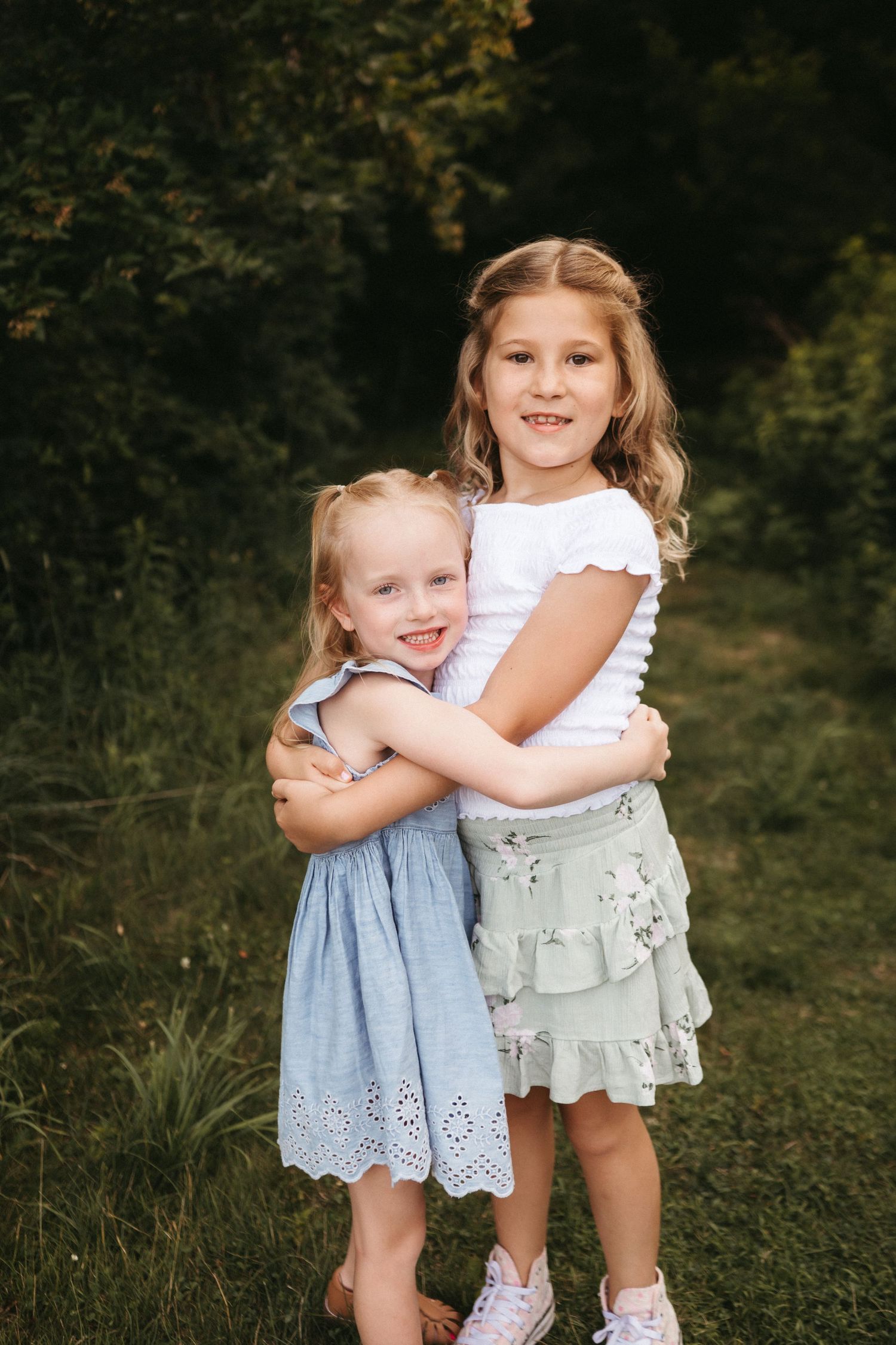 Two young children wearing summer dresses embrace while standing in a grassy outdoor setting.