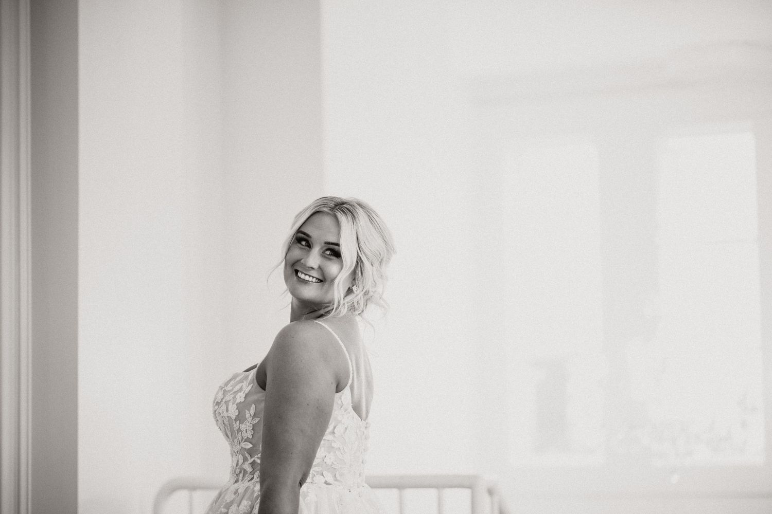 A radiant bride in a white lace gown poses by a bright window.