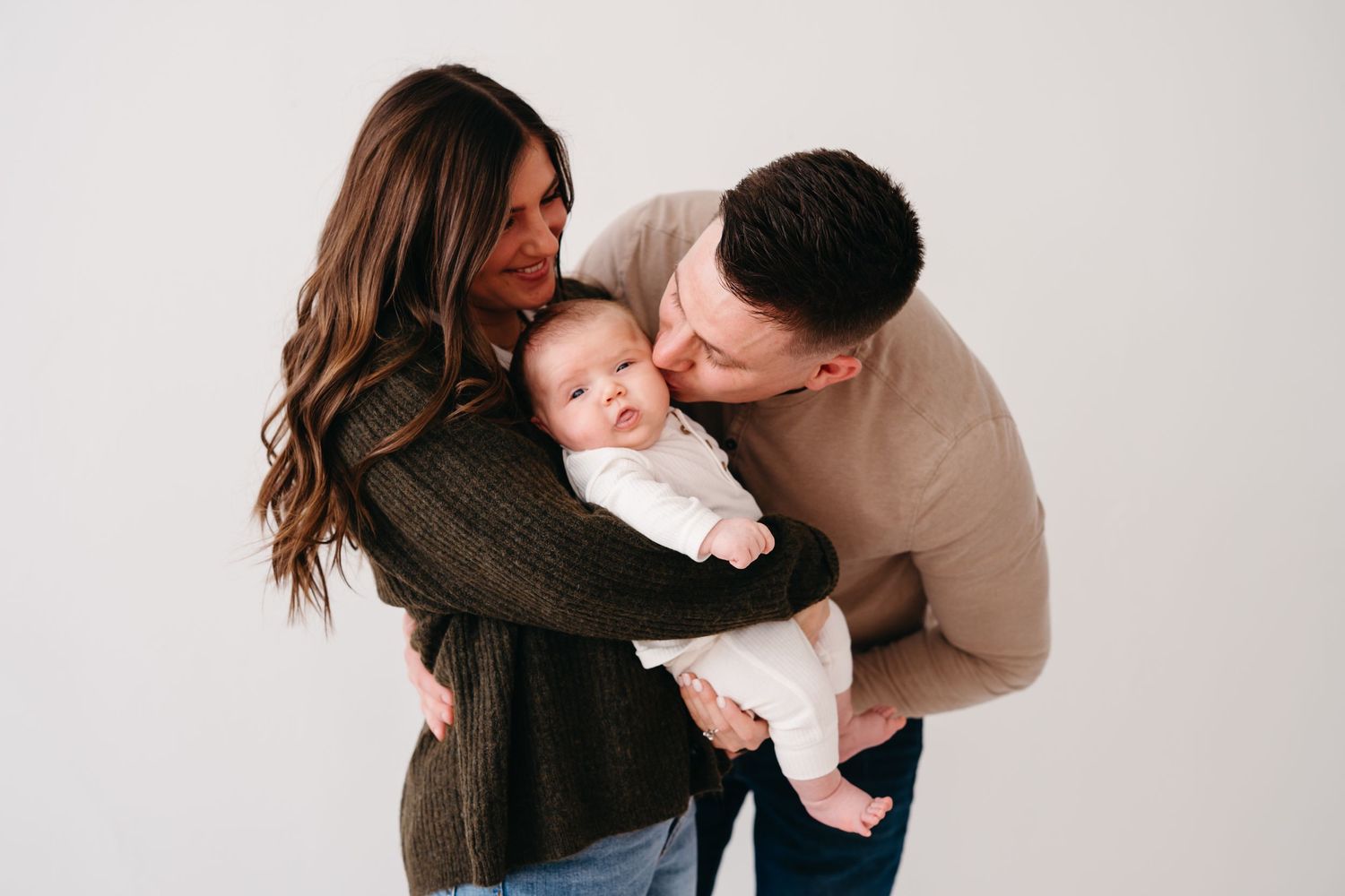 A couple holding and kissing their baby while standing against a white wall.