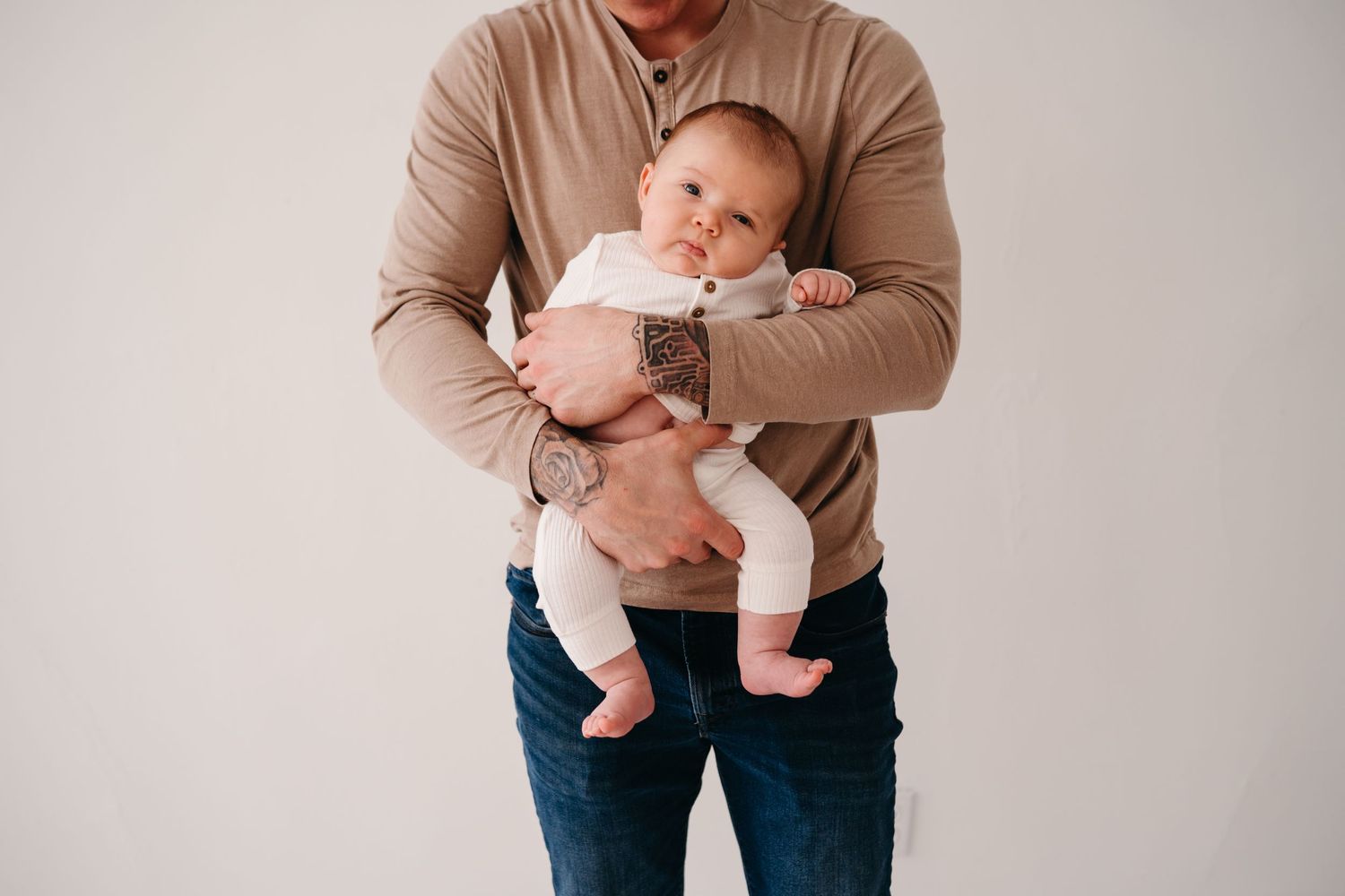 Parent tenderly holding a baby wearing white clothing against neutral background.