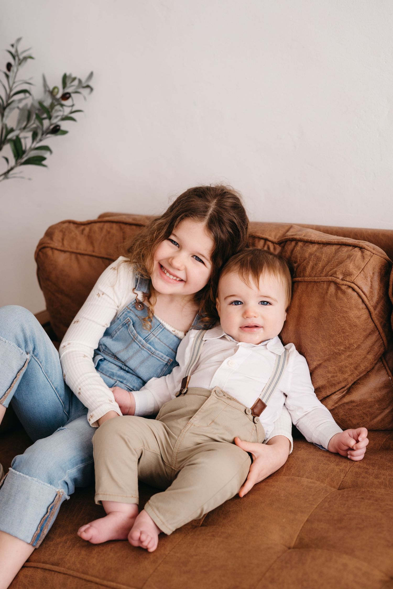 Two young children sit together on a brown leather couch, wearing casual clothes and sharing happy smiles.