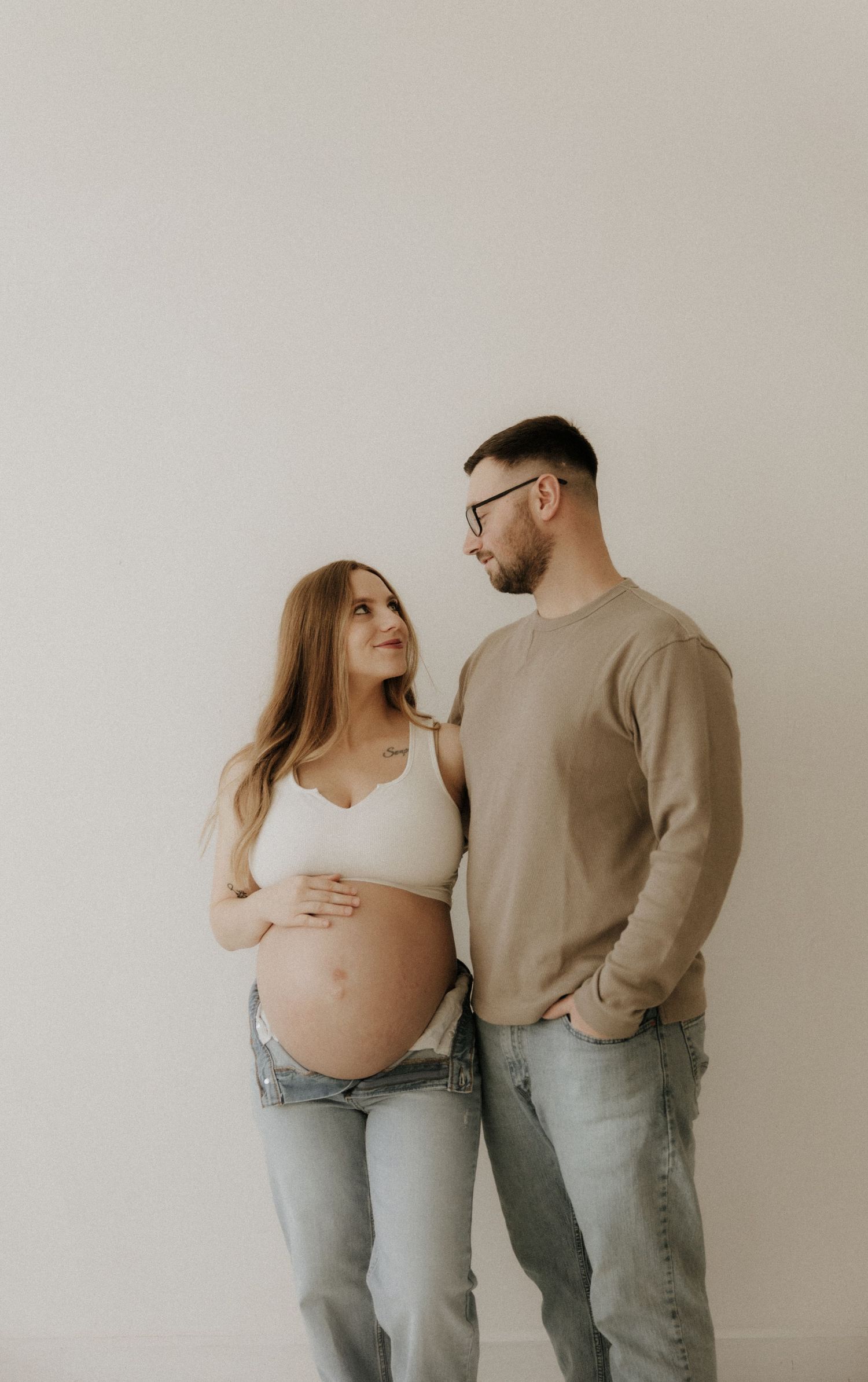 A couple in matching beige tops and jeans pose together during a maternity photoshoot against a neutral wall.