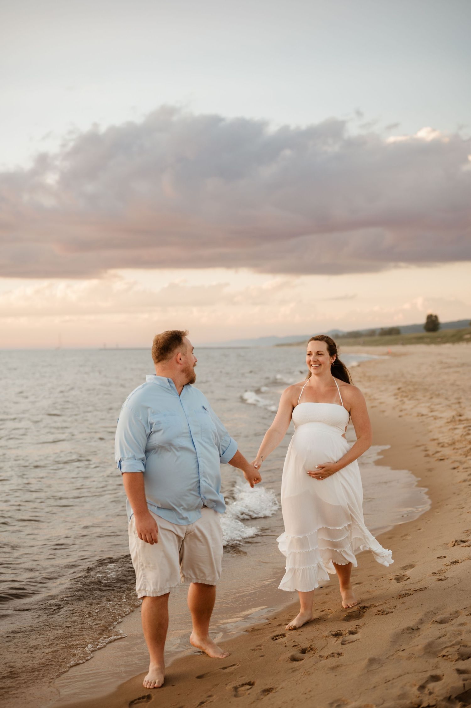 A couple walks hand in hand along a sandy beach at sunset with soft clouds and ocean waves in the background.