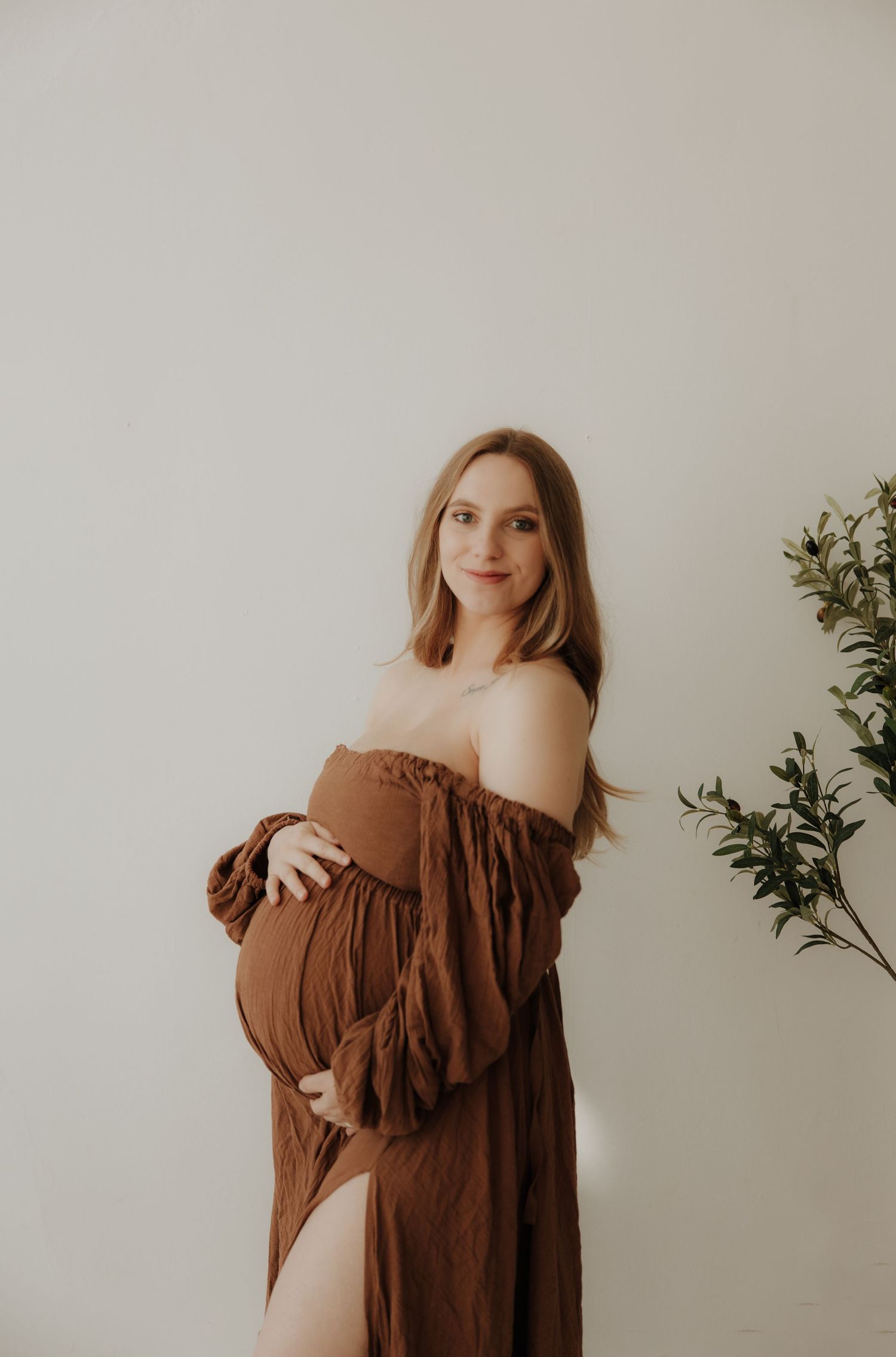 Romantic brown dress maternity photos with greenery accent in background.
