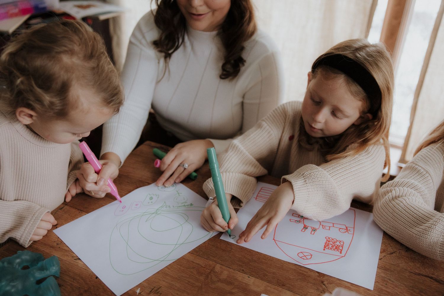 Children and adult work on drawings together at a wooden table.
