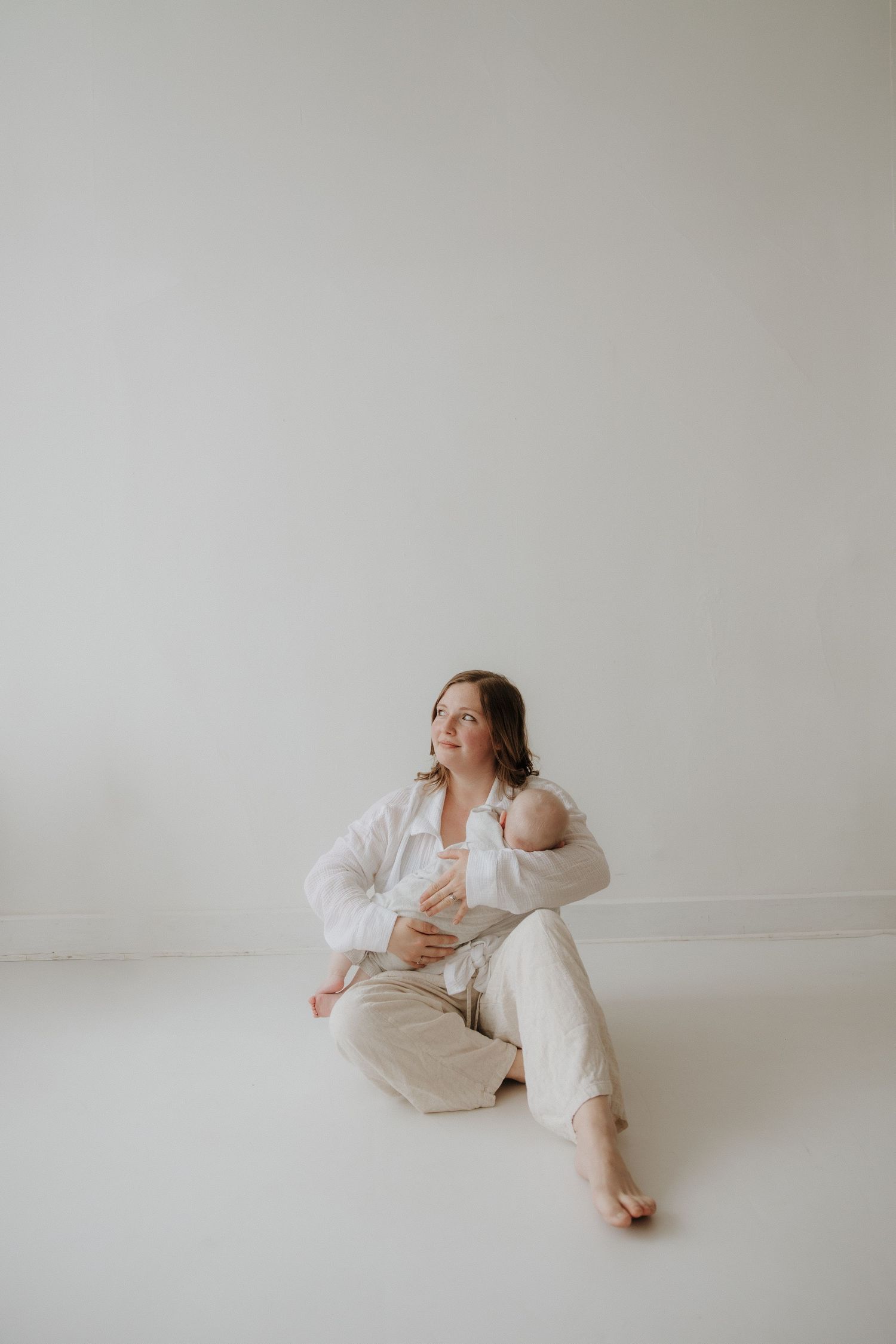 A mother sits barefoot on a white floor cradling her newborn baby while wearing a white linen outfit in a bright studio.