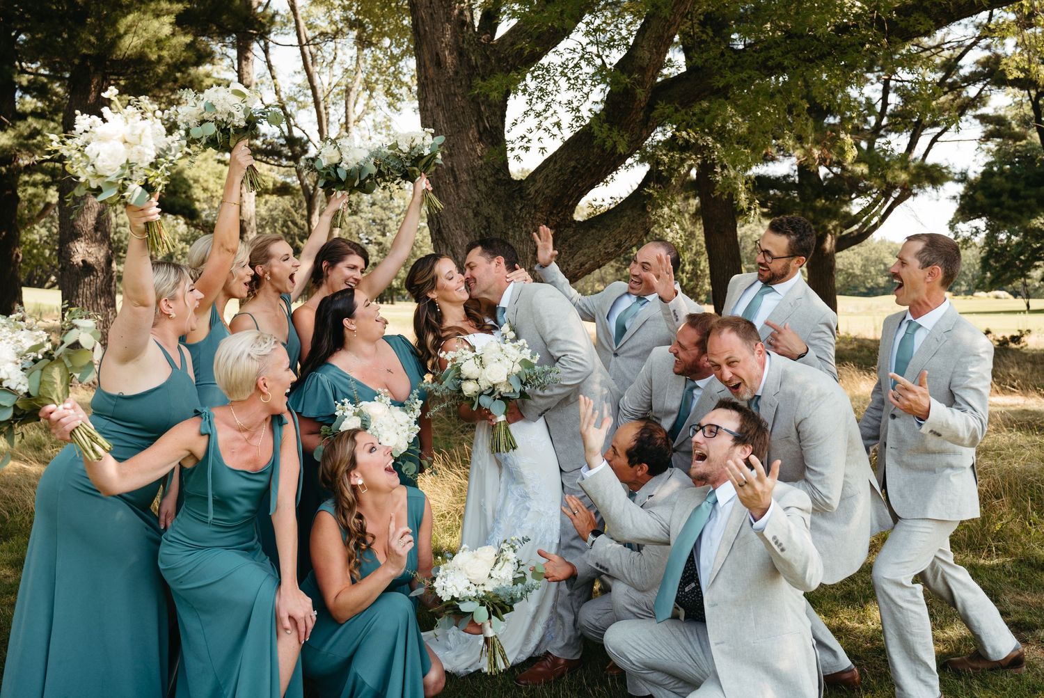 Wedding party in teal and gray attire shares laughs during outdoor photo session.