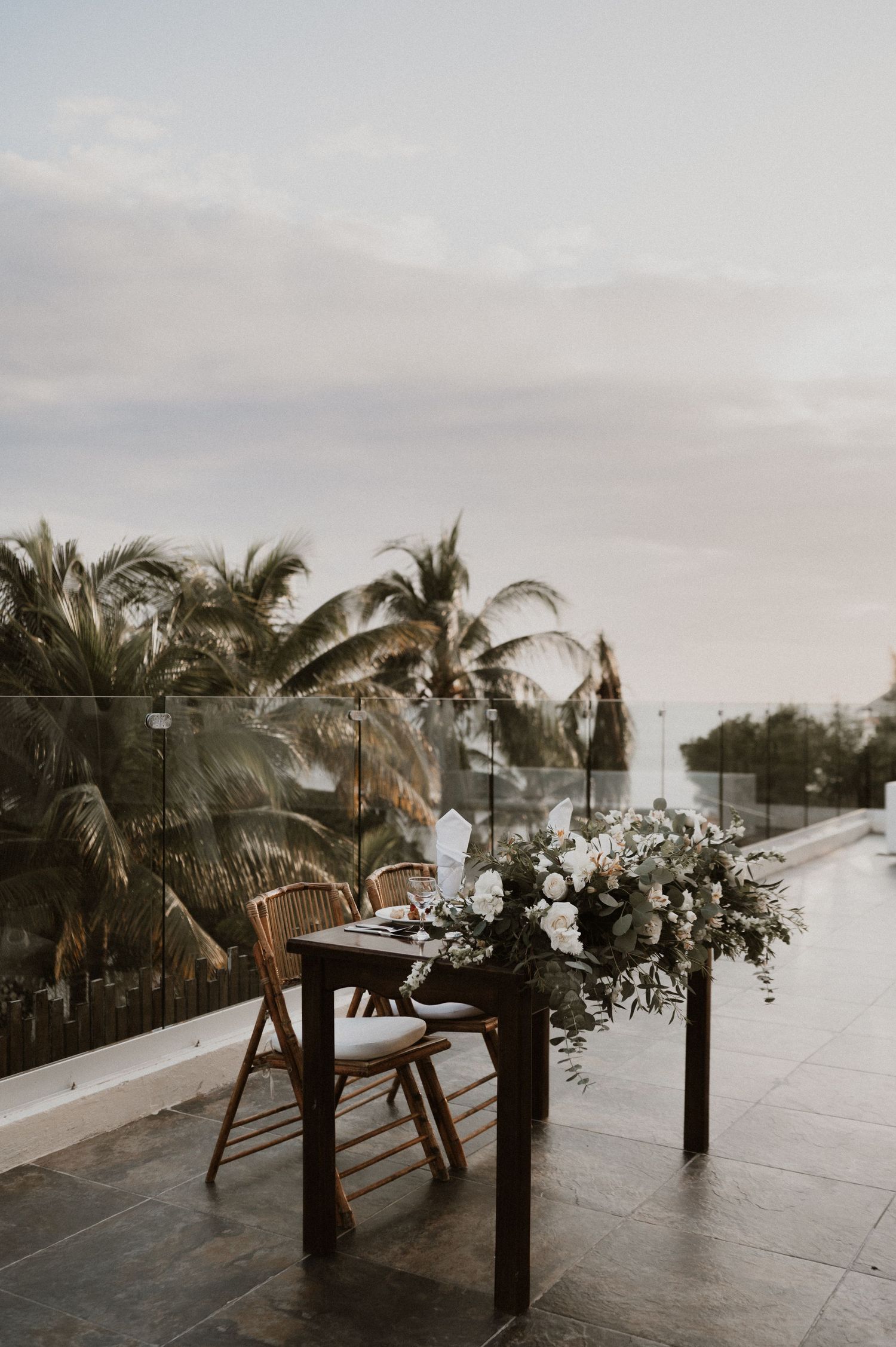 Elegant outdoor wedding setup with wooden chairs and floral arrangements against palm trees at sunset.