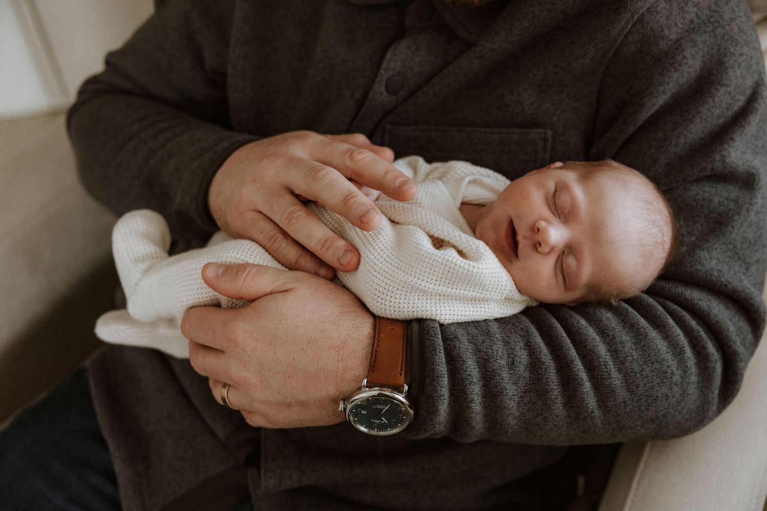 A newborn baby wearing white sleeps peacefully in someone's arms wearing a gray sweater and wristwatch.