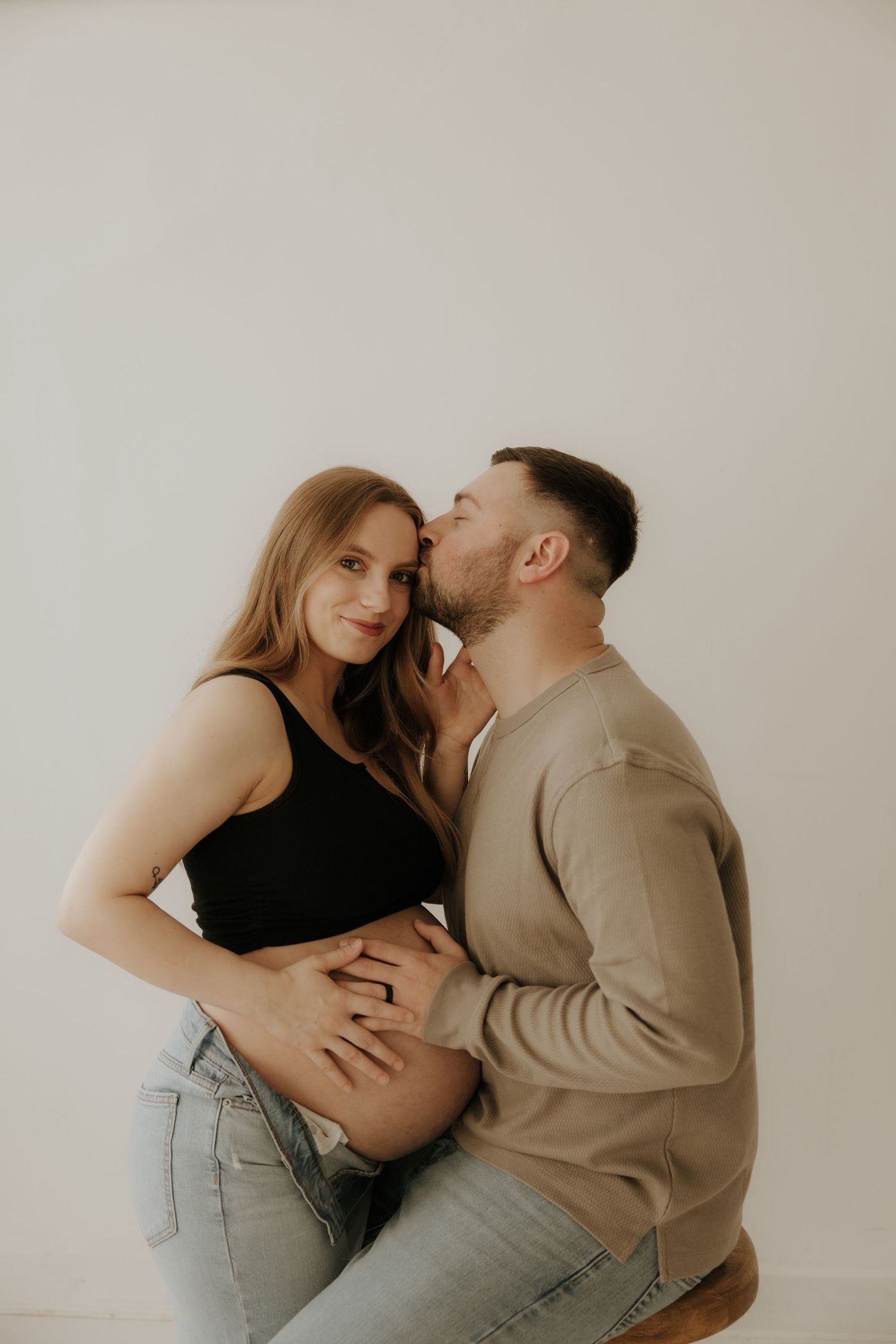 Intimate maternity photoshoot featuring couple in casual attire against neutral background.