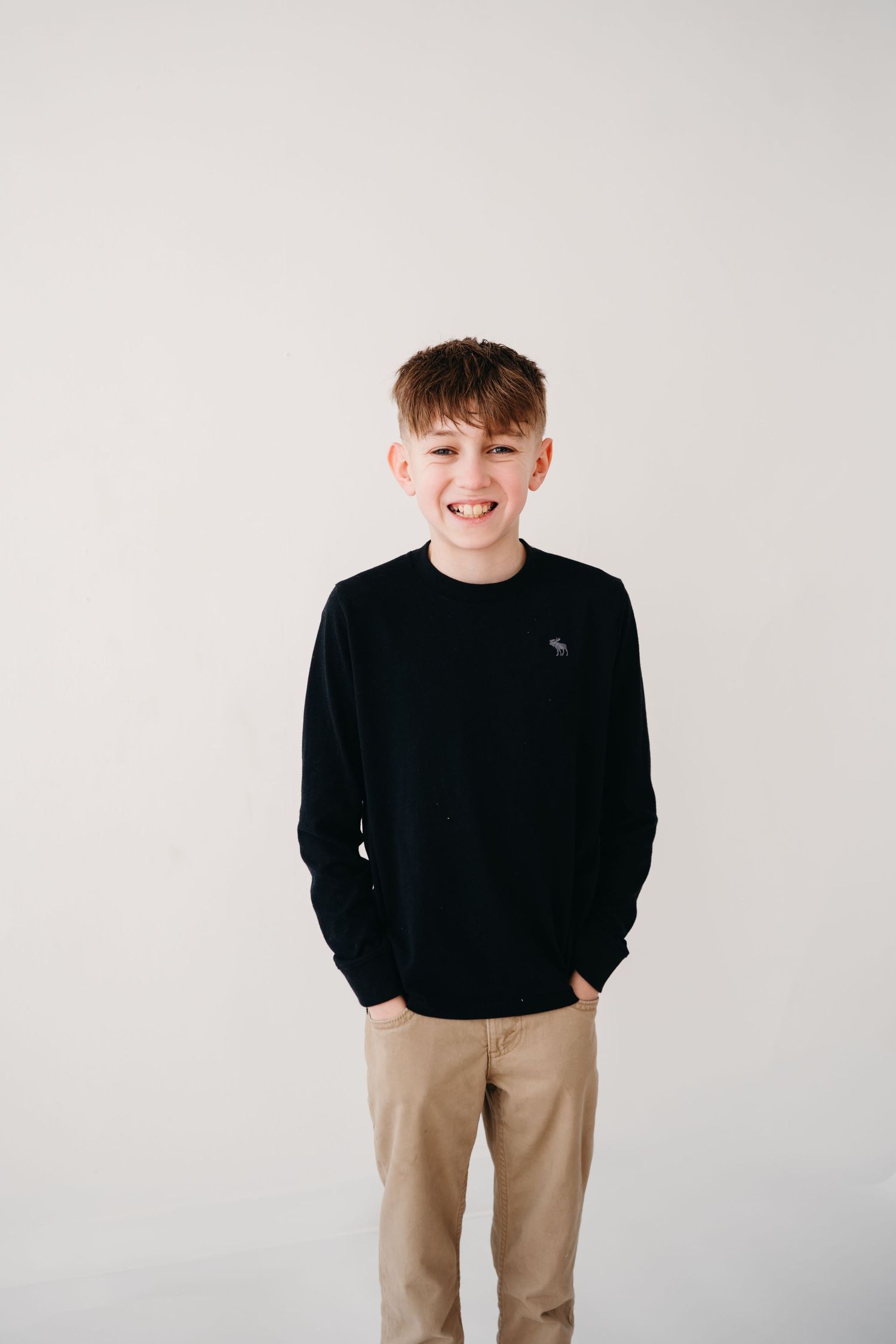 A young person in a black sweater and khaki pants stands smiling against a white background.