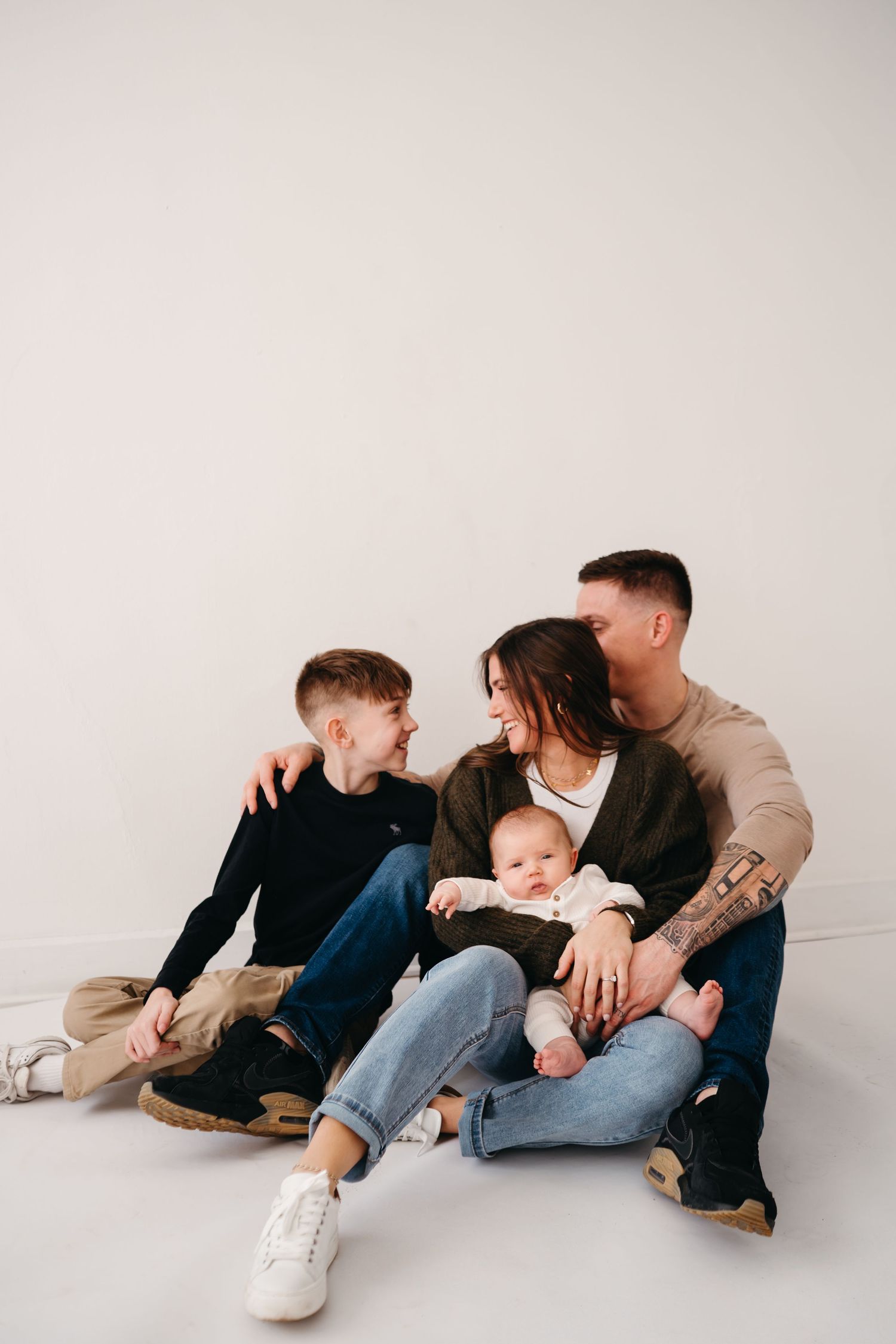 Family sitting together on floor against white wall wearing casual clothing and cuddling.