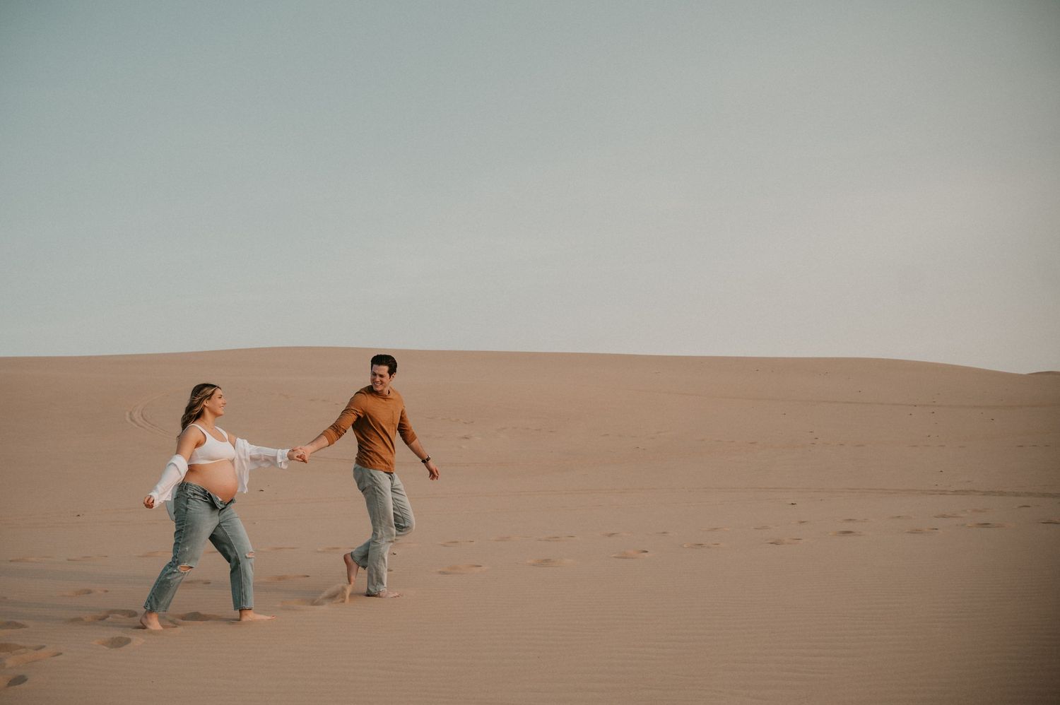 A couple holds hands while walking across golden sand dunes during sunset.