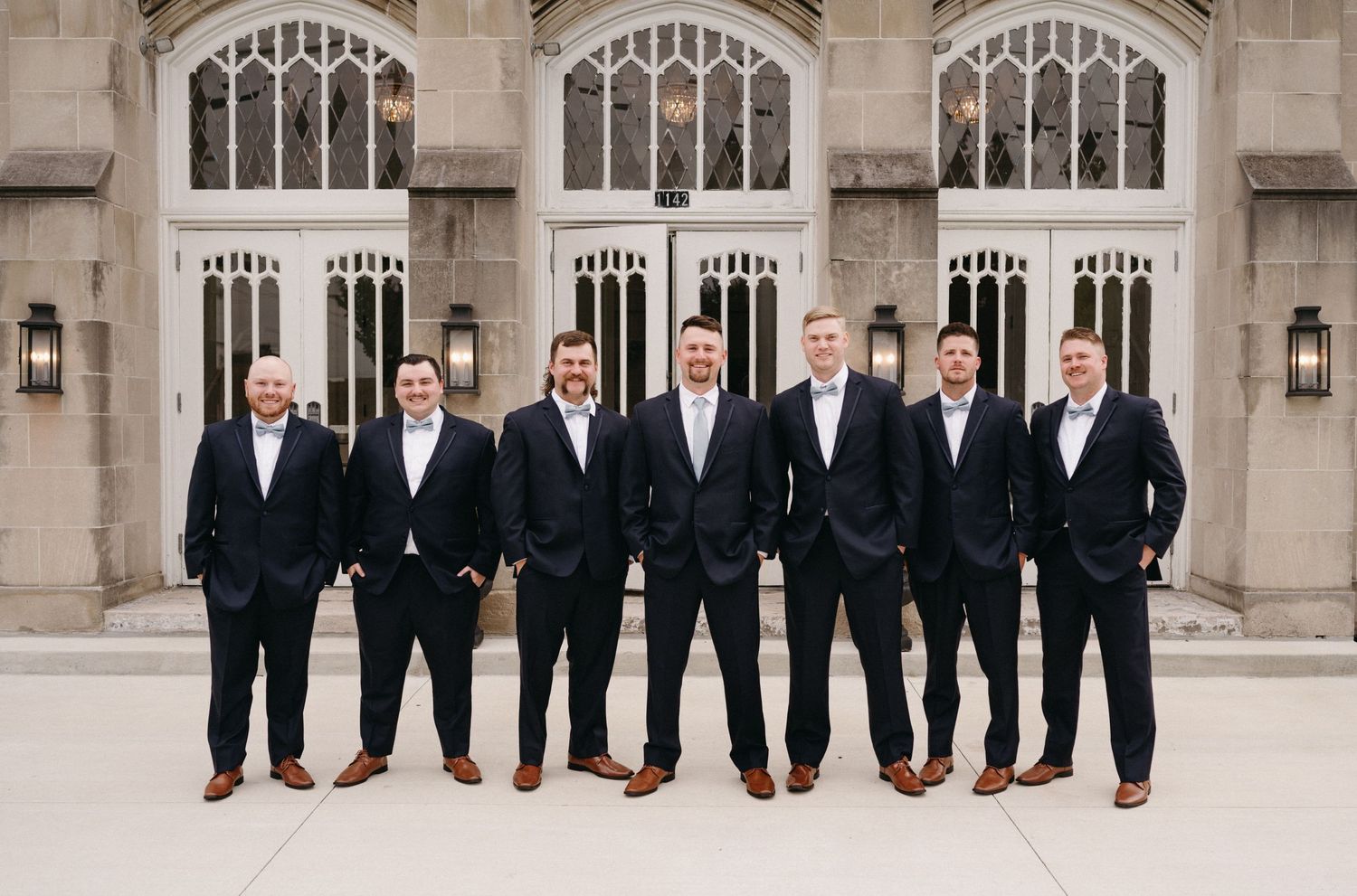 Groomsmen in matching dark suits and brown shoes pose in front of ornate church architecture.