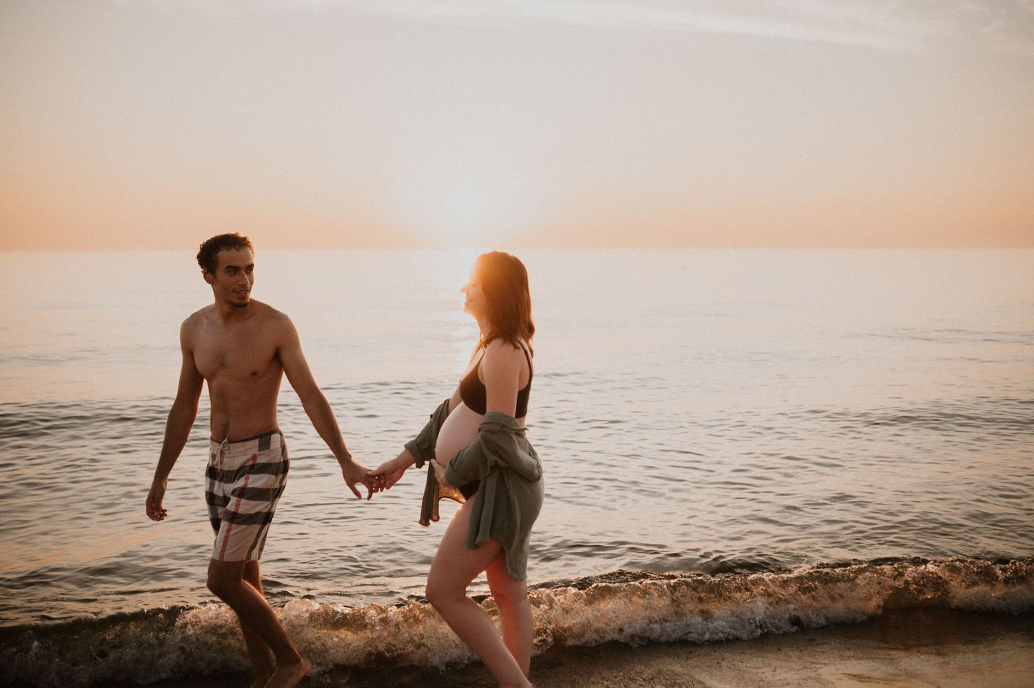 Couple holding hands while walking along beach shoreline at sunset.