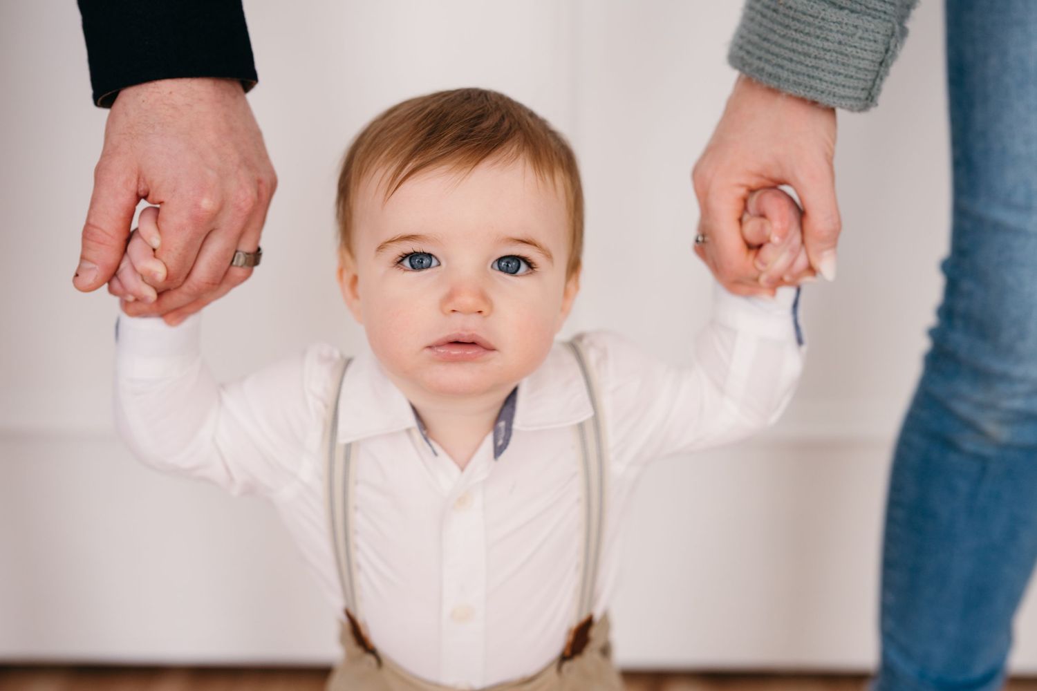 Small child in white outfit and suspenders holding hands with parents.