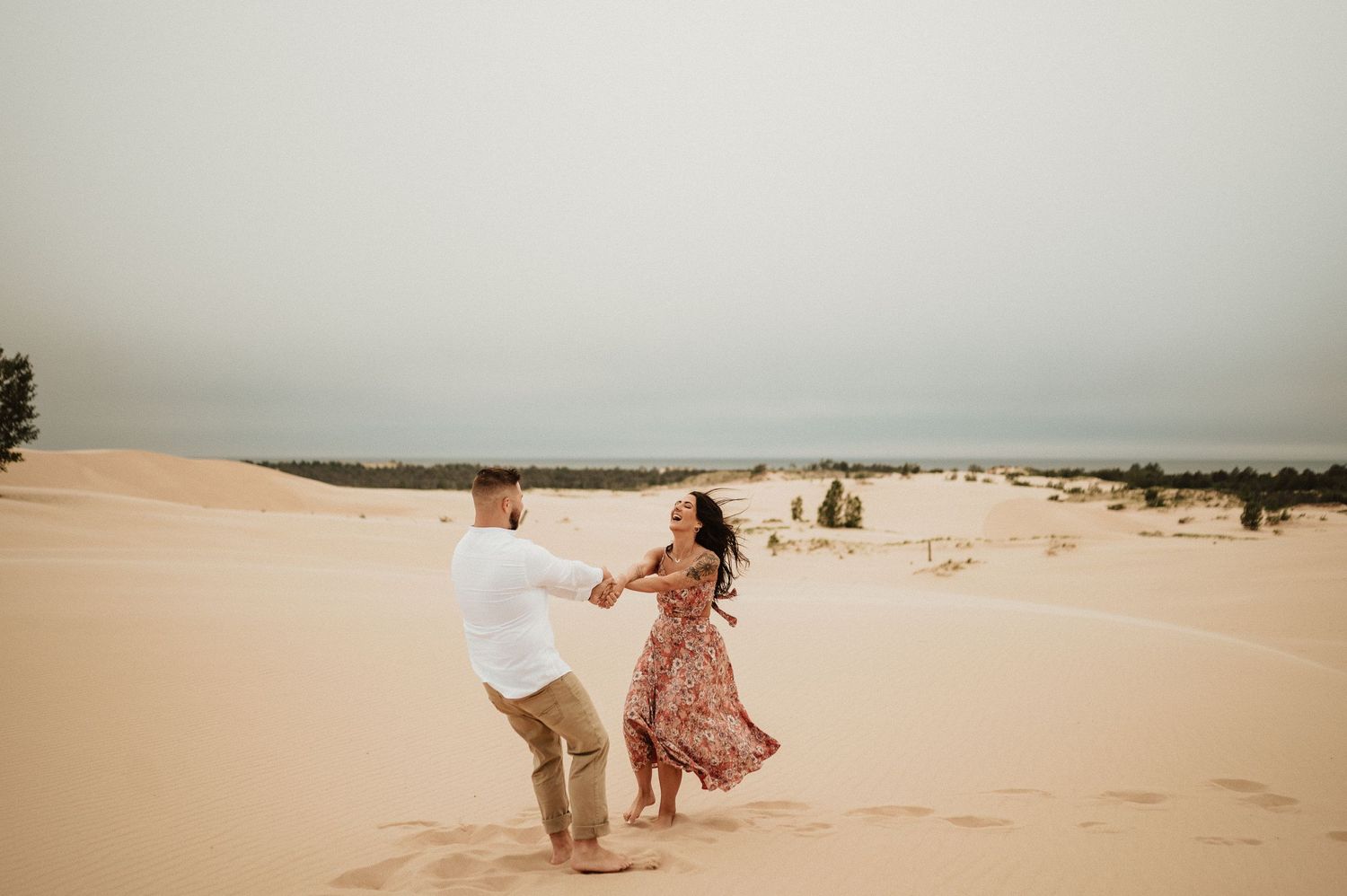 A joyful couple dances across desert sand dunes wearing coordinated white and floral outfits.