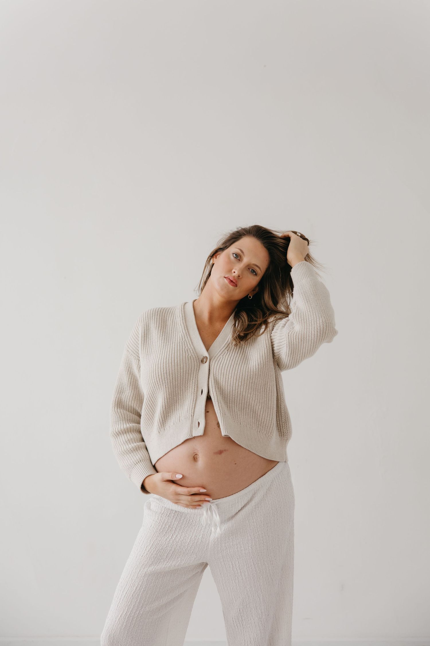 Pregnant woman in cream knit cardigan and pants posing with hand in hair on white background.