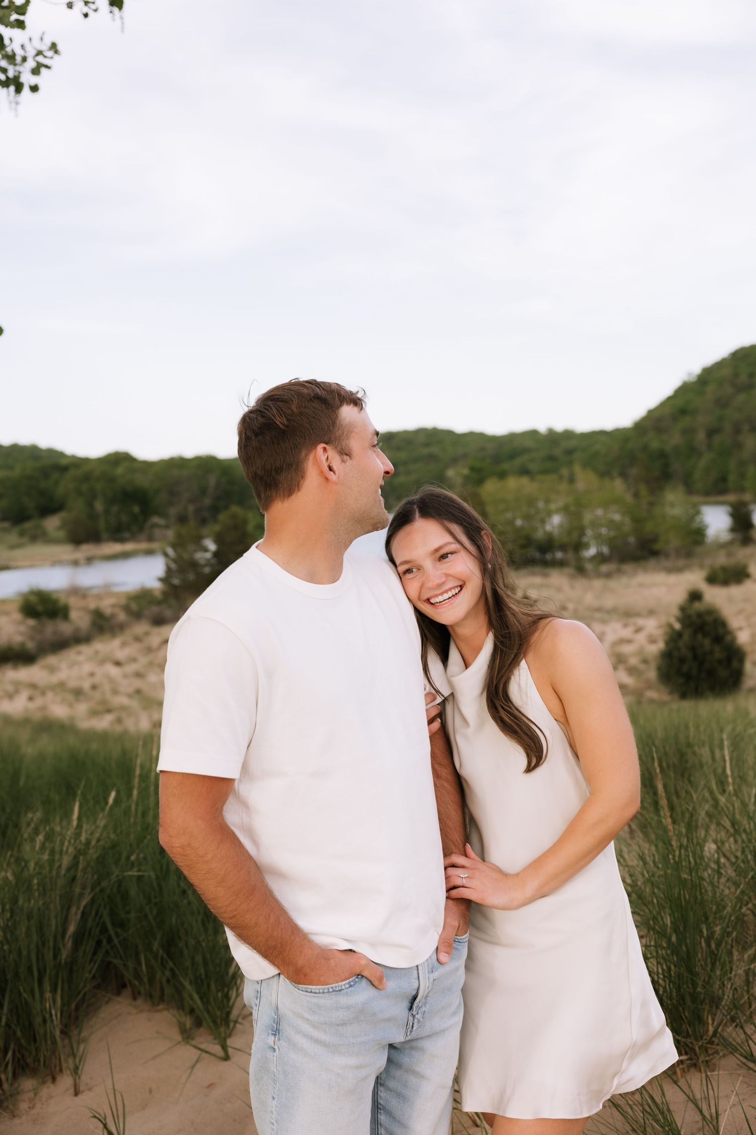 Happy couple posing on sandy beach dunes, woman in white dress leaning on mans shoulder.
