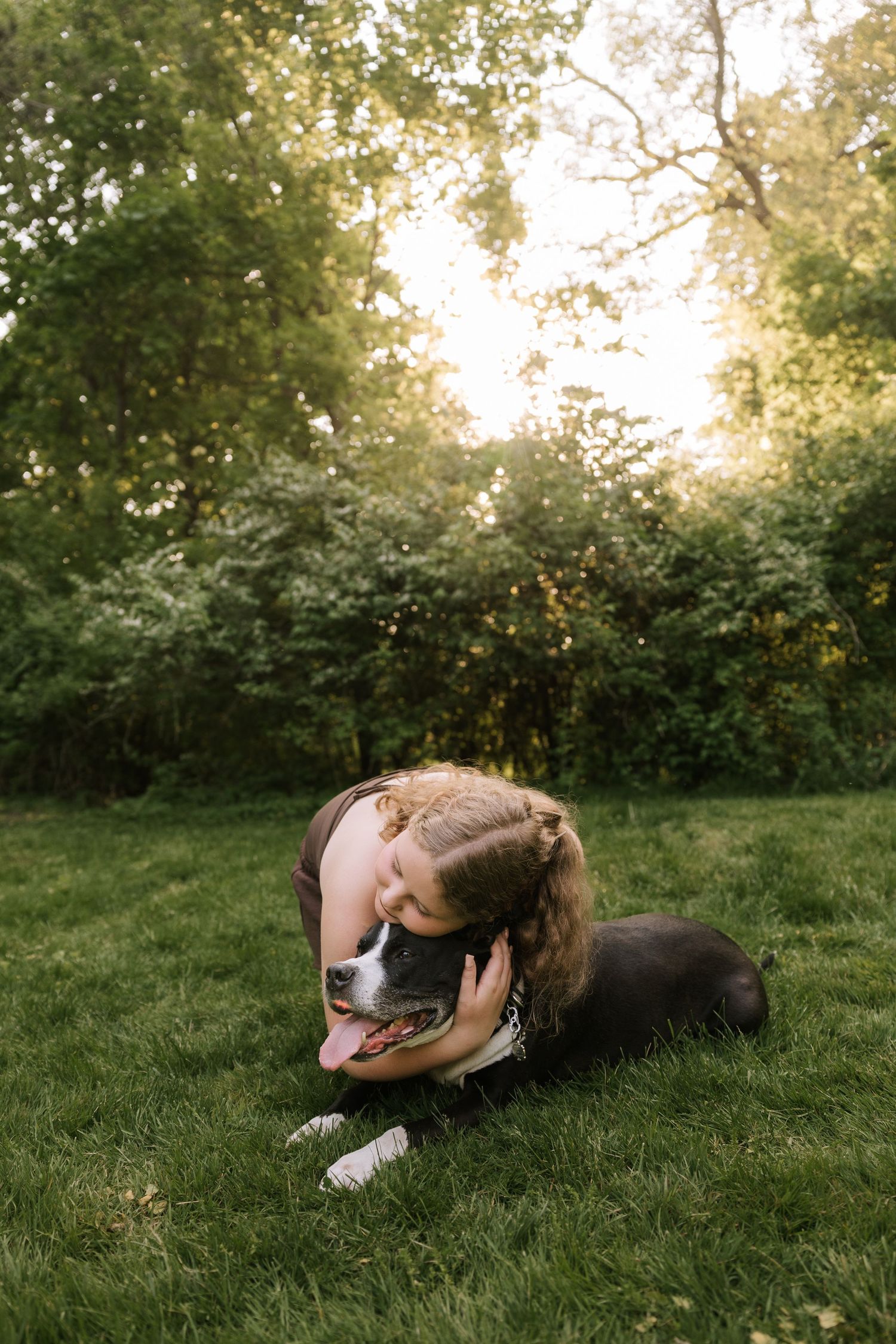 Young girl hugging black and white dog while sitting on green grass in sunny backyard.